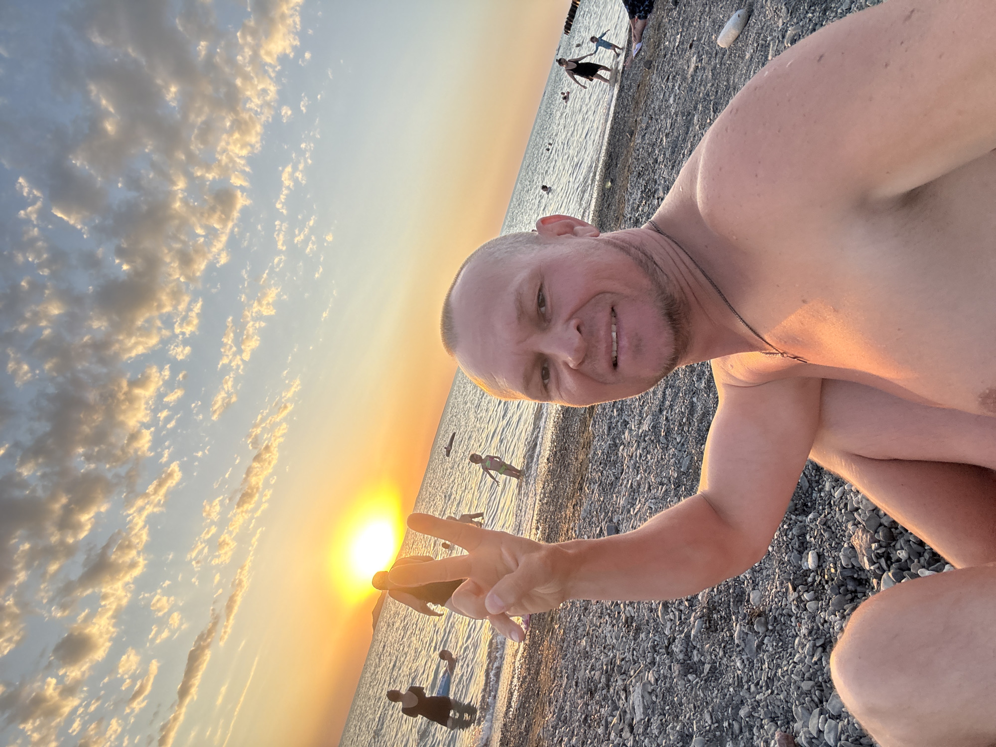 The image depicts a man taking a selfie on a pebble beach during sunset. He is shirtless, smiling, and holding up two fingers in a peace sign. Several other people, including adults and children, are visible in the water behind him, some wading and others swimming. The sun is low on the horizon, casting a warm, golden light across the scene, and the sky is filled with scattered clouds. The background features the sea meeting the shore. There is no indication of the image being located in the city of Urdzhar, Kazakhstan.