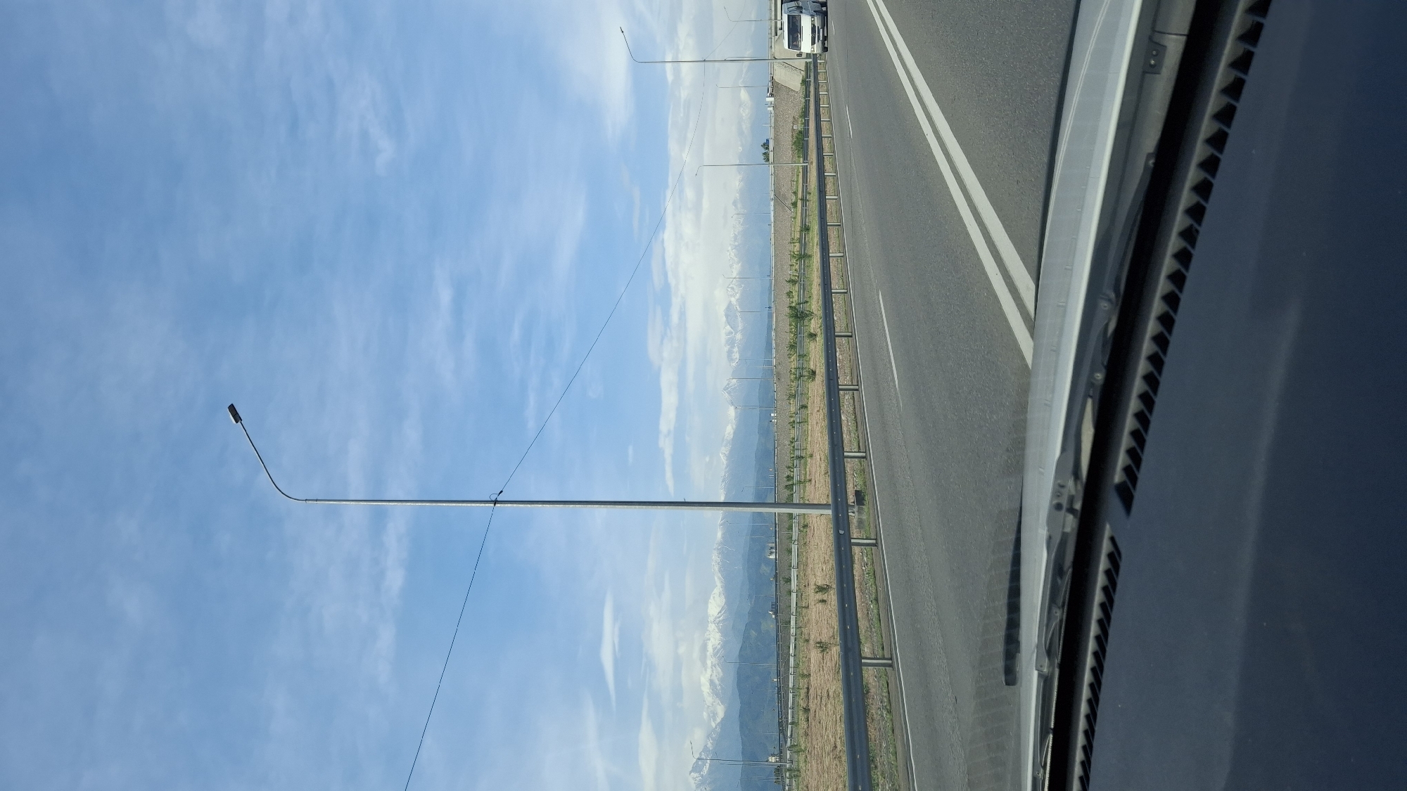 The image shows a view from inside a vehicle, likely a car, driving on a highway. The highway is divided by white lines and has a metal guardrail along the side. Beyond the guardrail is a field of dry grass and low vegetation. In the distance, a mountain range is visible with some snow-capped peaks. A series of streetlights line the side of the road. The sky is blue with some clouds. The location is possibly near Панфилово, Kazakhstan based on contextual information. There is no indication of people, interactions, or actions. The emphasis is on the road and the natural landscape in the background.