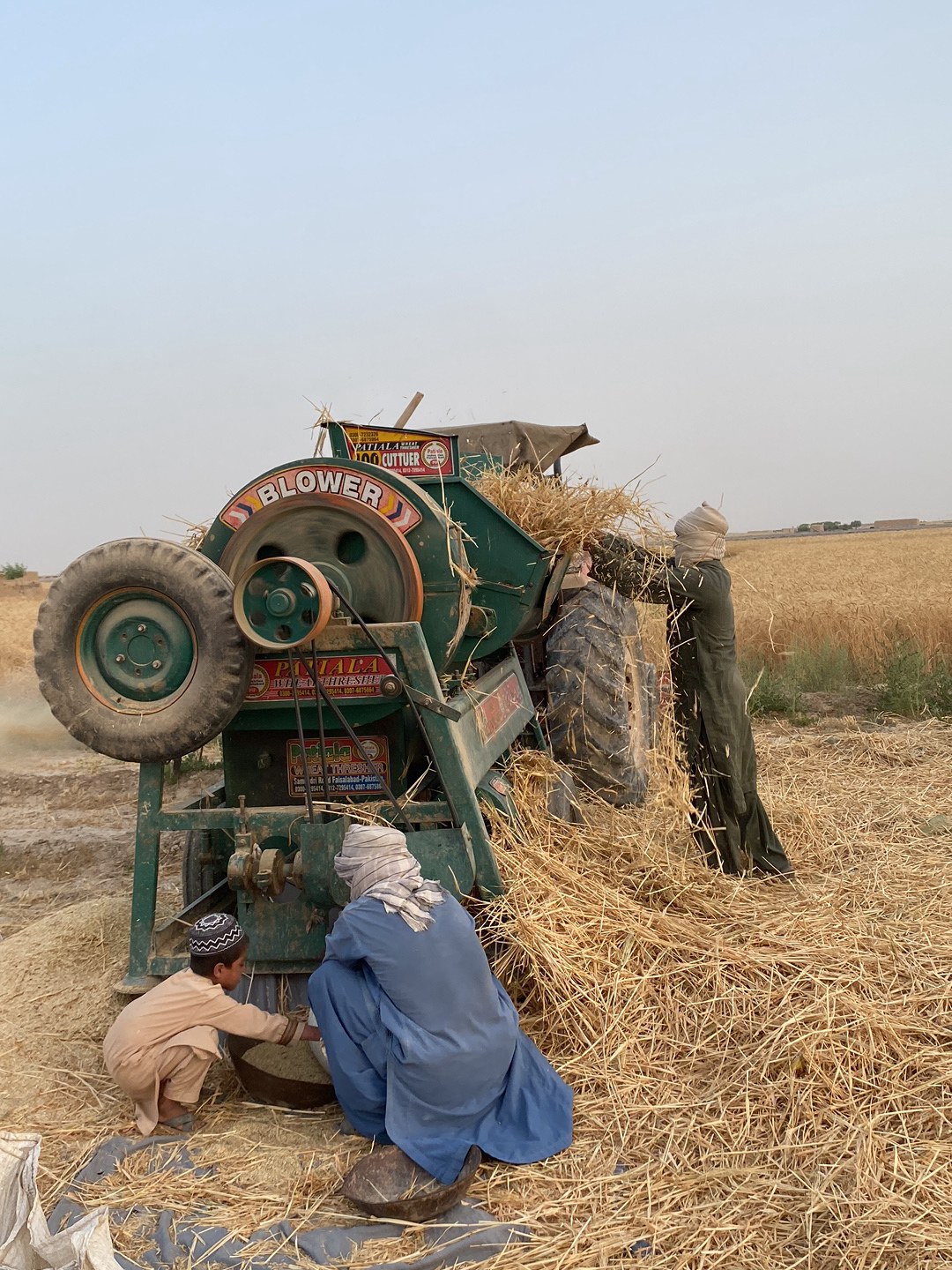 In an outdoor rural scene, likely in Alaqadari Daman, Afghanistan, a group of three people are working with a green agricultural machine. The machine is labeled "BLOWER" with additional text on the sides and a sign above that reads "SUPER 100 CUTTER". A child wearing a patterned cap and light-colored clothing is squatting near the front of the machine. An adult wearing a blue garment and a turban is squatting beside the child. A third person wearing a long dark garment and a turban is standing near the back of the machine, feeding straw into the top of the machine. Straw is scattered around the base of the machine. The environment consists of a field of dry vegetation under a light sky.