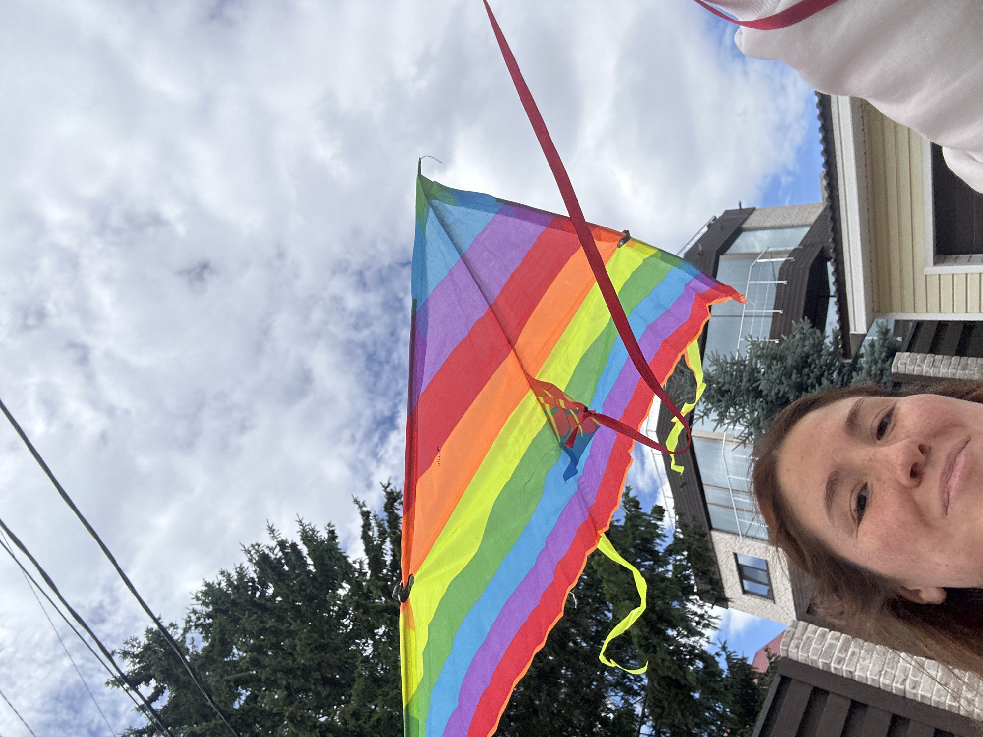 A young woman with reddish-brown hair and freckles is pictured from the chest up, smiling slightly. She is outdoors with a bright rainbow-colored kite dominating the upper portion of the image. The kite has red, orange, yellow, green, blue, and purple stripes. The woman is positioned in the lower left quadrant of the frame. Behind her, a modern house with large windows and a balcony is visible, partially obscured by evergreen trees. Electrical wires stretch across the sky. The sky is cloudy with patches of blue. The setting appears to be a residential area in Nizhny Novgorod, Russia, given the architectural style of the house and the presence of coniferous trees.