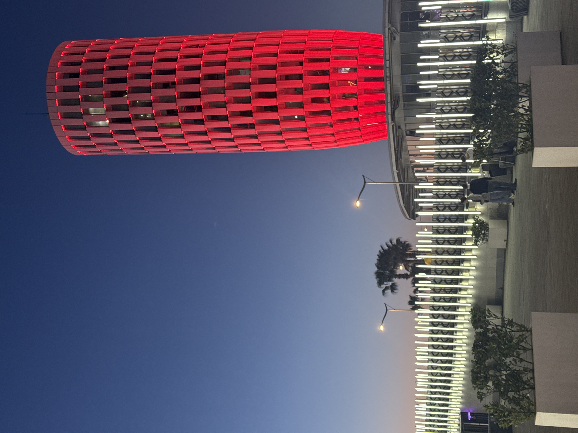The image shows a section of the city of Batumi, Georgia at dusk. A cylindrical building with a lattice-like red exterior stands out against the gradient sky transitioning from deep blue to a lighter hue near the horizon. Adjacent to this structure is a taller building with horizontal white illuminated bars, possibly lights, running across its facade. Green vegetation, including trees and plants, are interspersed along the base of the taller building. A couple of streetlights are visible casting a warm glow. People are walking along the building's base, suggesting an urban environment.