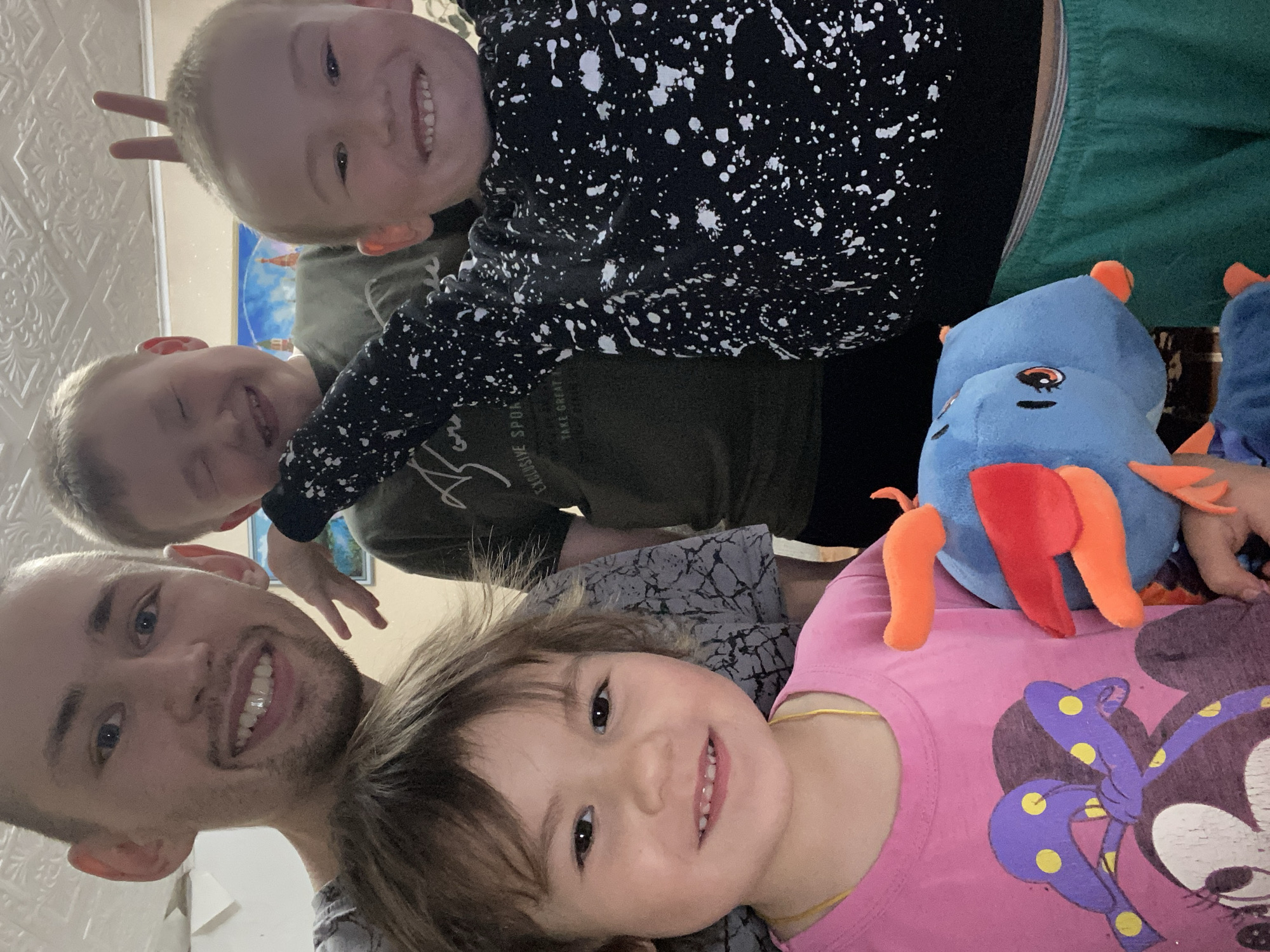 A selfie shows a fair-skinned man with light hair and a beard, smiling at the bottom left. Above him are three children with fair skin and light hair, also smiling. One child holds up two fingers behind another child's head. A stuffed blue animal with orange and red accents lies next to the child in the pink shirt. The environment shows a room with wallpaper, and the scene appears to be indoors, possibly in Bely Klyuch, Russia.