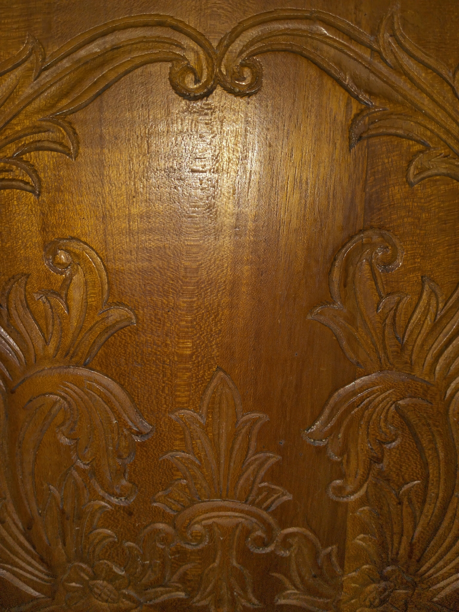 This is a close-up, vertical shot of ornate, carved wood. The wood has a rich, warm brown color and a visible grain pattern. The carving features swirling, floral-like motifs with leaf and scroll elements. The carving is detailed and appears to be a decorative panel or part of a larger piece of furniture. The lighting creates highlights and shadows that accentuate the depth and texture of the carving. There are no people, actions, or interactions depicted. The location in the city of Payagaboh, Indonesia, is not ascertainable from the image itself.