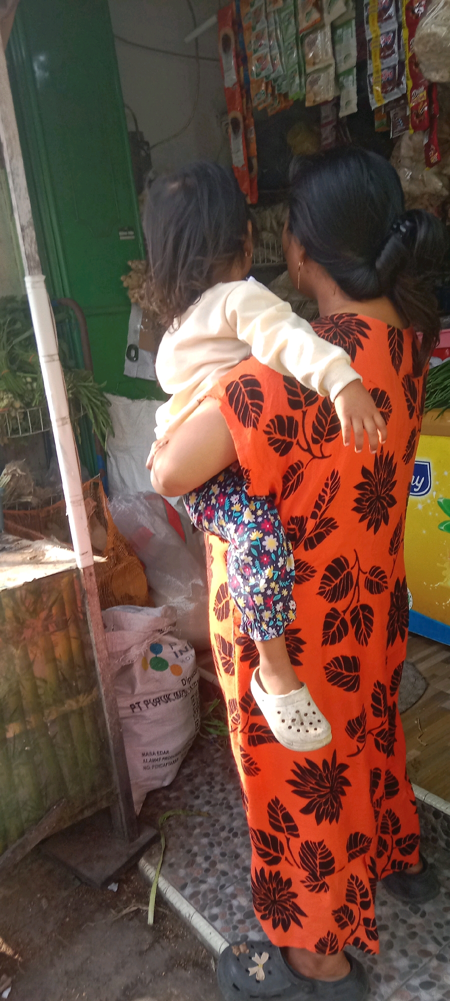 In Kras, Indonesia, a woman with dark hair pulled back in a bun is carrying a young child who is facing away from the camera. The woman is wearing a bright orange dress with a large black floral pattern. The child is wearing a light-colored long-sleeved shirt and patterned leggings with blue, white, and red flowers. The child has on a white Croc-style shoe on their right foot. The woman is wearing dark gray Croc-style shoes. They are standing on a surface composed of small, dark, and light gray pebbles. Behind the woman, shelves are stocked with various packaged goods, some appearing to be food items or snacks. To the left, a green wooden door is visible, and a vertical white pole with horizontal markings is present. A large white bag with text and graphics, possibly containing fertilizer or grains, is on the ground in the lower left quadrant of the image. The overall setting appears to be an outdoor market stall or a small shop.