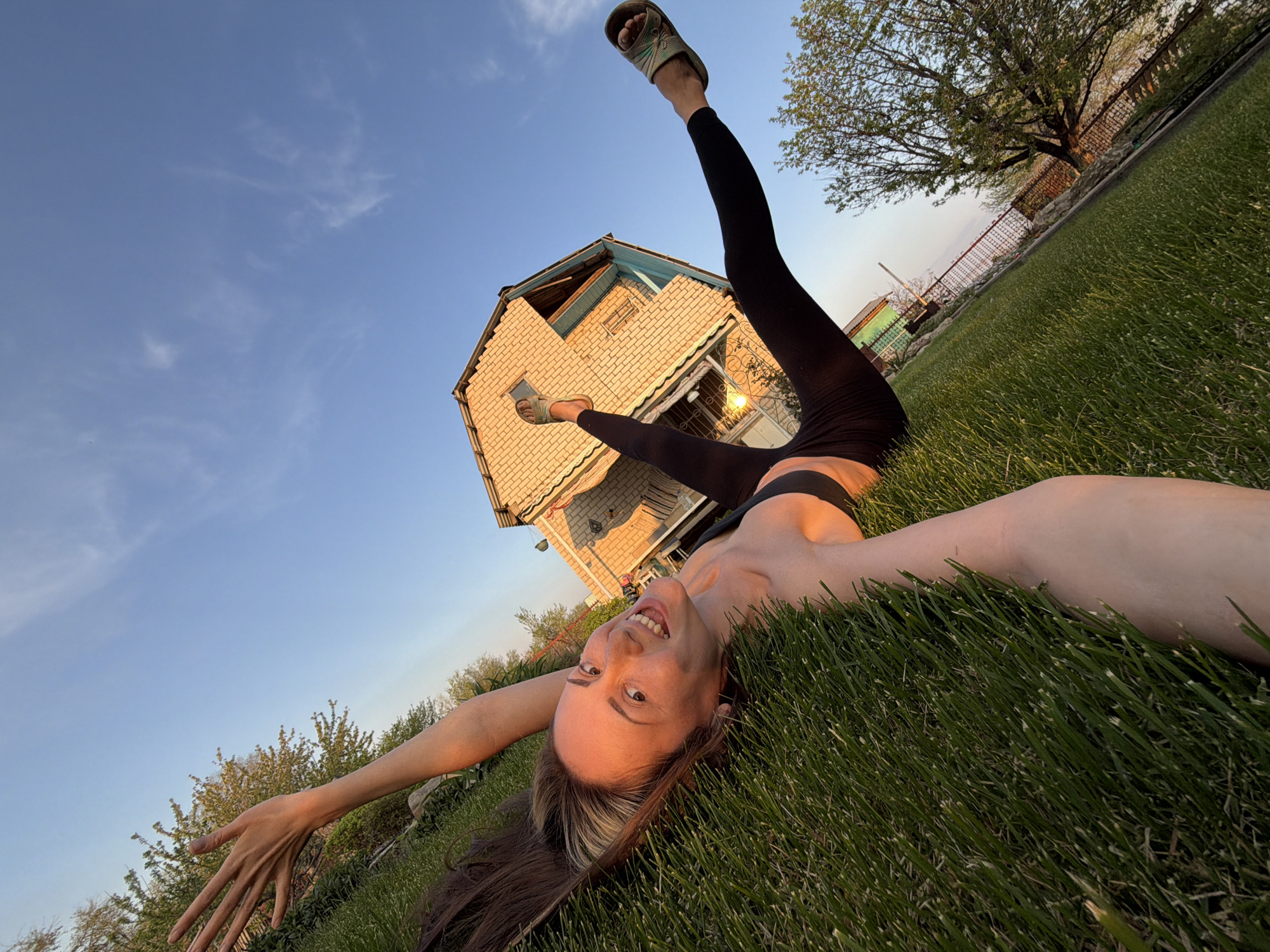 A young woman lies upside down on a lawn in Kamyshin, Russia. She wears black leggings and a black sports bra. Her hair is brown with a blonde streak. She is smiling and her arms are outstretched. Her legs are raised towards the sky. She is wearing green sandals on her feet. Behind her is a two-story brick house with a blue roof and a tree. The sky is blue with some clouds. The photo is taken from a low angle.