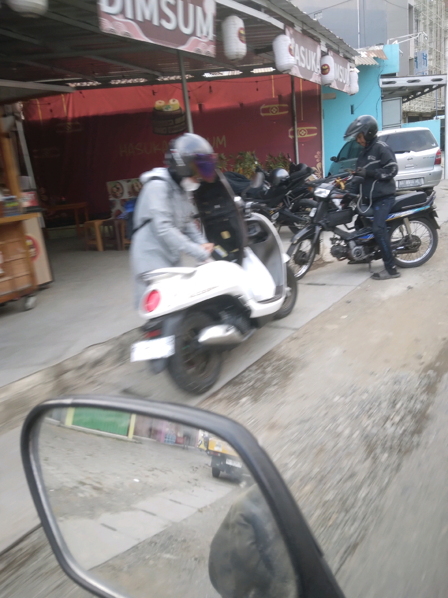 In Sokaraja, Indonesia, on a blurred street with asphalt and gravel, a person in a grey jacket and helmet rides a white scooter. To the right, another person wearing a black jacket and helmet is seated on a black motorcycle. In the background, a silver car with license plate AA 8916 HL is parked. A red awning with "DIMSUM" written on it is visible above a building. The scene appears to be in front of a food establishment.