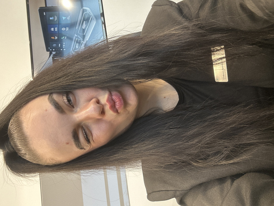 An adult female is captured in a close-up, head-and-torso shot within an indoor commercial setting. She possesses long, dark hair, partially styled in a high ponytail with strands falling over her shoulders and frame. Her left eye is closed in a wink, while her lips are pursed in a 'kissing' gesture. She is attired in a dark-colored blazer over a black top. A translucent name tag is clipped to her right chest (viewer's left), displaying the Russian text "София Дрозд" (Sofia Drozd), with "Администратор" (Administrator) visible below, partially obscured.
In the background, a large, wall-mounted flat-screen monitor is visible behind her right shoulder (viewer's left). The monitor displays an automotive infotainment system interface, featuring a grid of icons for functions such as "Music," "Navigation," "Phone," and "Settings," along with a graphical representation of vehicle climate controls or a gear selector area. This interface is consistent with infotainment systems found in Chery brand vehicles. Faint branding, likely "CHERY" or "CHIMA," is discernible on the lower bezel of the monitor.
Based on the Russian language present on the name tag and the specific automotive branding on the screen display, the scene is identified as occurring within a Chery car dealership or showroom in Saint Petersburg, Russia.