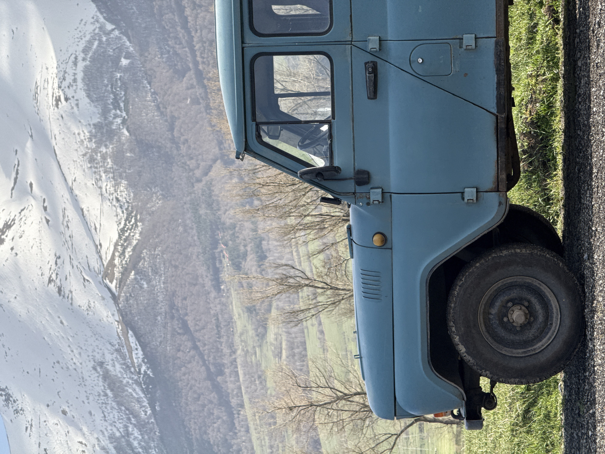 The image captures a light blue vehicle, likely a truck, parked alongside a road in Margahovit, Armenia. The tire of the vehicle is visible, set against a backdrop of green grass and an asphalt road. In the distance, a mountainous landscape is visible, partially covered in snow, juxtaposed against valleys and sparse trees, indicating a rural or mountainous setting.