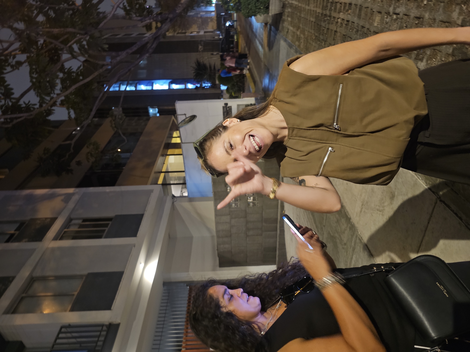The image depicts two women standing outside in what appears to be an urban setting at night. One woman, with shoulder-length hair and wearing a brown sleeveless top with zipper details, is smiling and making a hand gesture. The other woman, who has curly hair and is wearing sunglasses and a black top, is looking at and using a smartphone. A black purse is visible near her arm. The background includes a building with visible windows, street lights, and a tree. There are also several people walking in the background. Based on the architectural style and street layout, the location could potentially be the city of Miraflores in Peru.