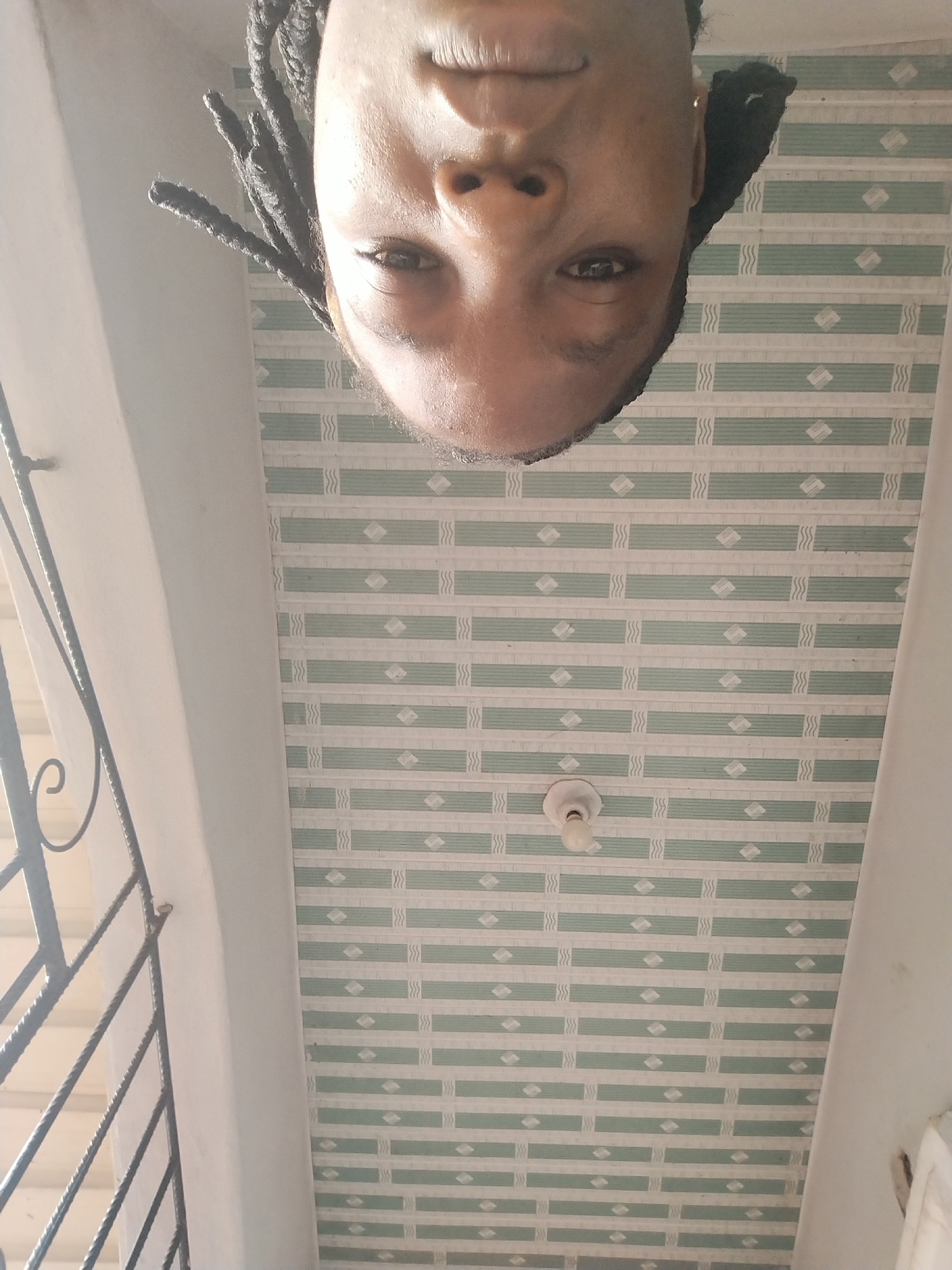 The image, taken from a low angle looking upwards, features a person's face upside down in the foreground. The individual has dark-colored dreadlocks and appears to be looking directly at the camera. Their skin tone is brown. Behind the face, the ceiling is visible, adorned with a repeating pattern of light green and white horizontal rectangular tiles. A simple white light fixture with a bare bulb is centrally located on the ceiling. To the left of the frame, a portion of a white wall and a black metal grate or railing is visible, suggesting an interior space with a window or balcony. The lighting appears to be natural, casting soft shadows. This scene is located in Isiehor, Nigeria.
