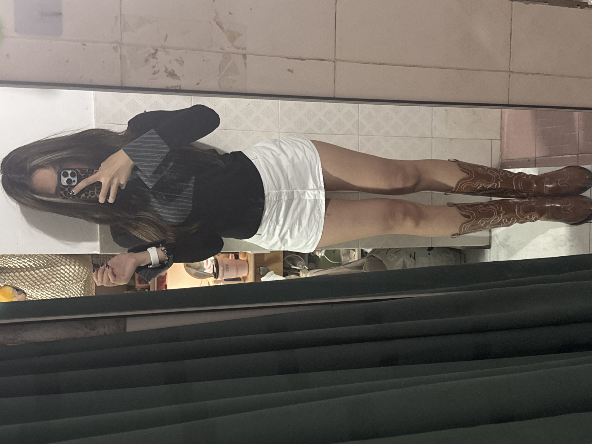 A woman is taking a mirror selfie in what appears to be an indoor setting. She is wearing a black blazer, a white skirt, and brown cowboy boots. She holds a phone with a leopard-print case in front of her face. The location seems to be a domestic space in Nan'an District, China, based on the visible items like a straw basket, and some domestic appliances in the background. The backdrop is tiled walls and a dark curtain.