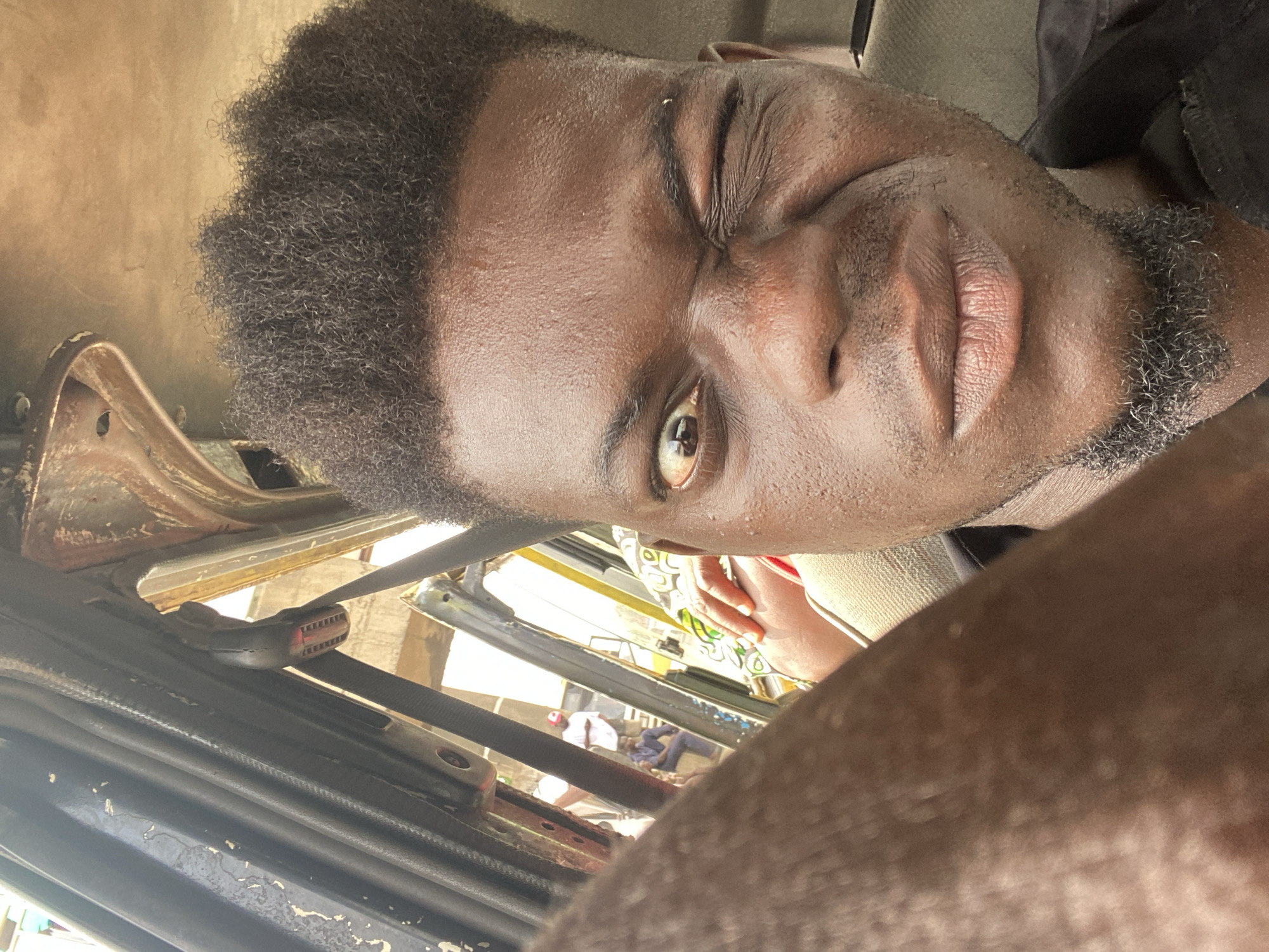 This media file features a close-up, upward-angled portrait of a man, rotated 90 degrees counter-clockwise. The man, of African descent, has dark skin, short, curly black hair, and some facial stubble around his chin and jawline. He looks directly at the camera with a playful expression, winking with his right eye, and his lips are slightly parted.
The setting appears to be inside a vehicle, likely a car or a commercial passenger vehicle, indicated by the visible seatbelt buckle and components of a door or window frame. Through the vehicle's opening, a blurred outdoor background is visible, showing parts of buildings or structures and indistinct figures, including one person wearing a red cap or head covering.
The image is captured in bright, natural light, suggesting it is daytime. The man's wink conveys a friendly or playful emotion. No visible text is present.