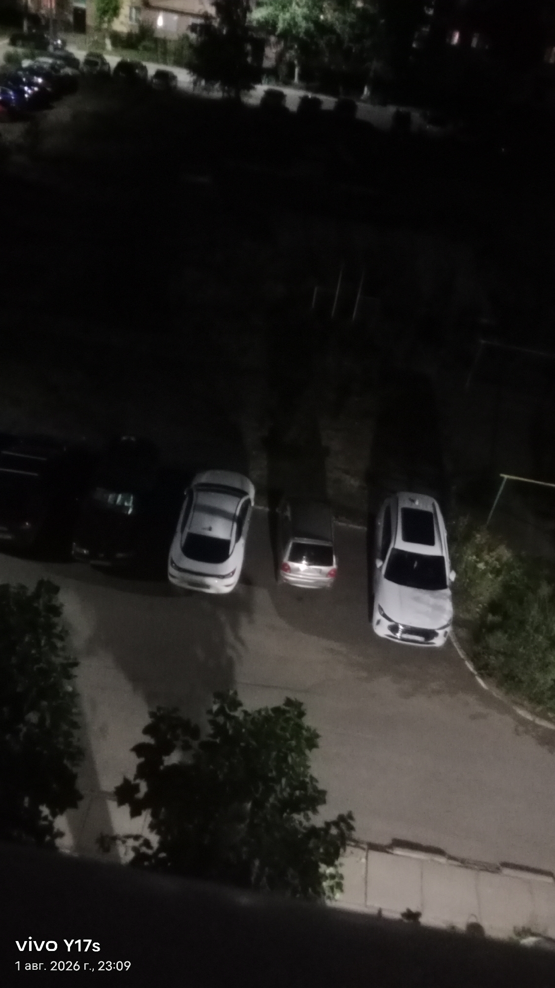 An elevated, nighttime view shows a street with several parked cars. The cars are white and silver, and some are dark. The street is paved with asphalt, and there is some greenery on the sides. The image is dated August 1, 2026, at 23:09. The location is indicated as Rudny, Kazakhstan, in the caption.