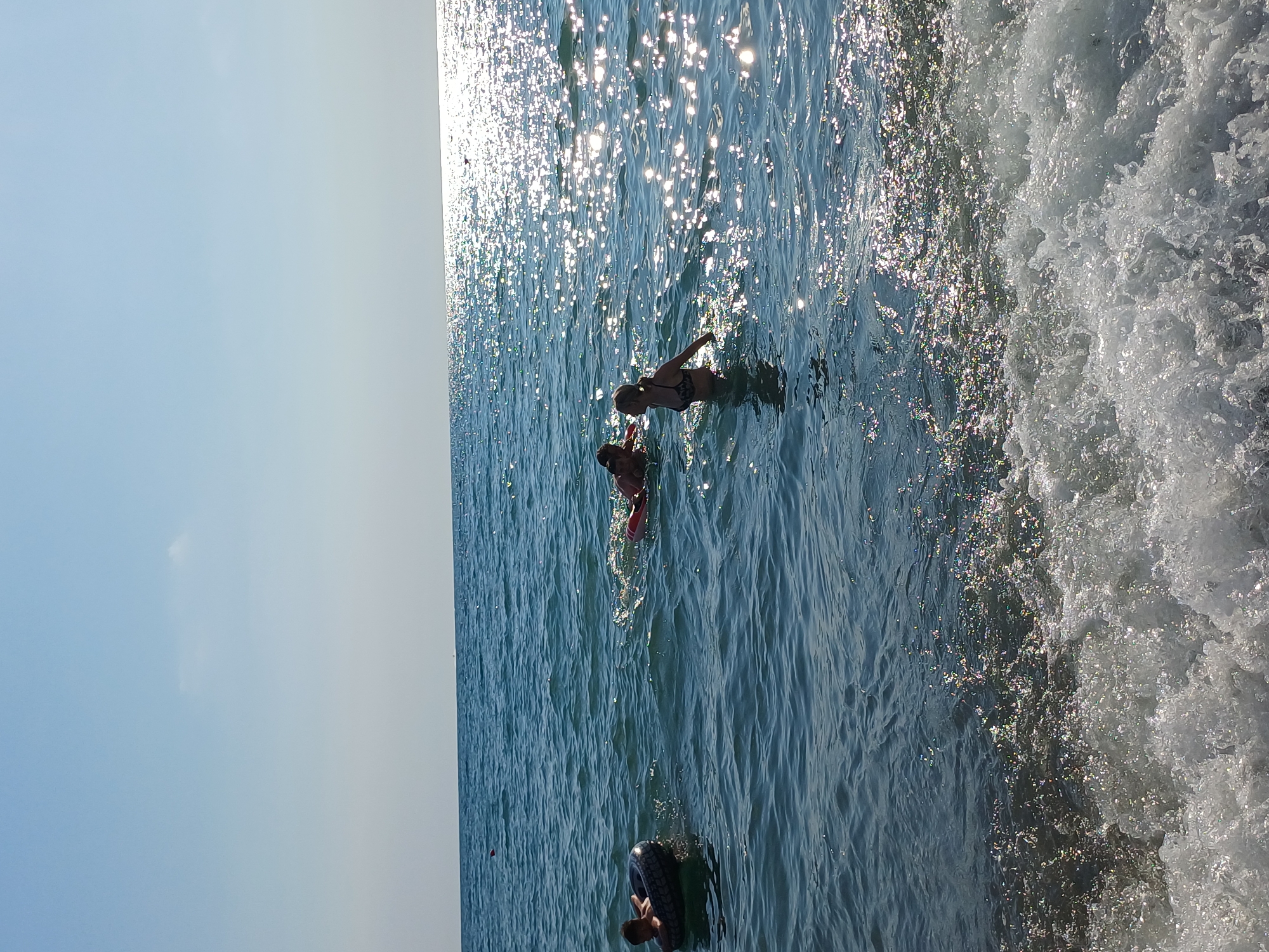 This image depicts a group of people enjoying a sunny day at the beach in Sochi, Russia. The scene is a bright, clear day with the sun glinting off the water, creating a sparkling effect. The water is a deep blue, with waves breaking on the right side of the frame, indicating a lively beach environment.
In the water, three individuals are visible. One person, wearing a bikini, is swimming and appears to be reaching out. Another person, partially submerged, is interacting with a red floatation device. Further down, another person is seen sitting in a tire inner tube. The activity appears to be recreational swimming and playing in the sea.
The overall impression is one of a pleasant summer day at the beach, with people engaging in typical beach activities. The bright sunlight suggests it is daytime, likely midday or afternoon, given the intensity of the reflections. No visible text is present in the image.
