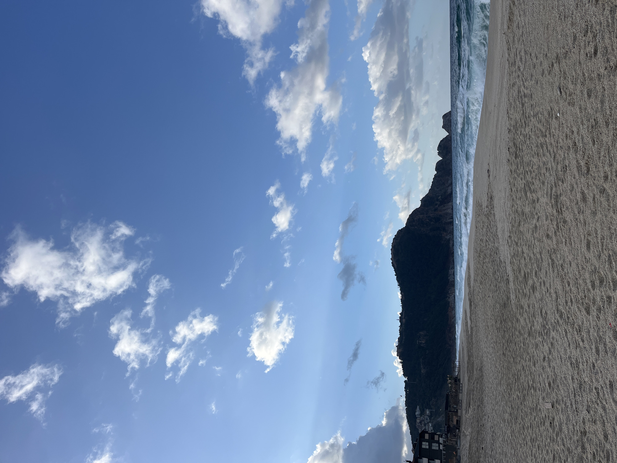 The image depicts a beach scene in Alanya, Turkey. The view includes a sandy beach in the foreground, with waves crashing on the shore. A dark, rocky mountain rises from the edge of the beach, with buildings visible on its lower slopes. Above, a blue sky is scattered with white clouds.