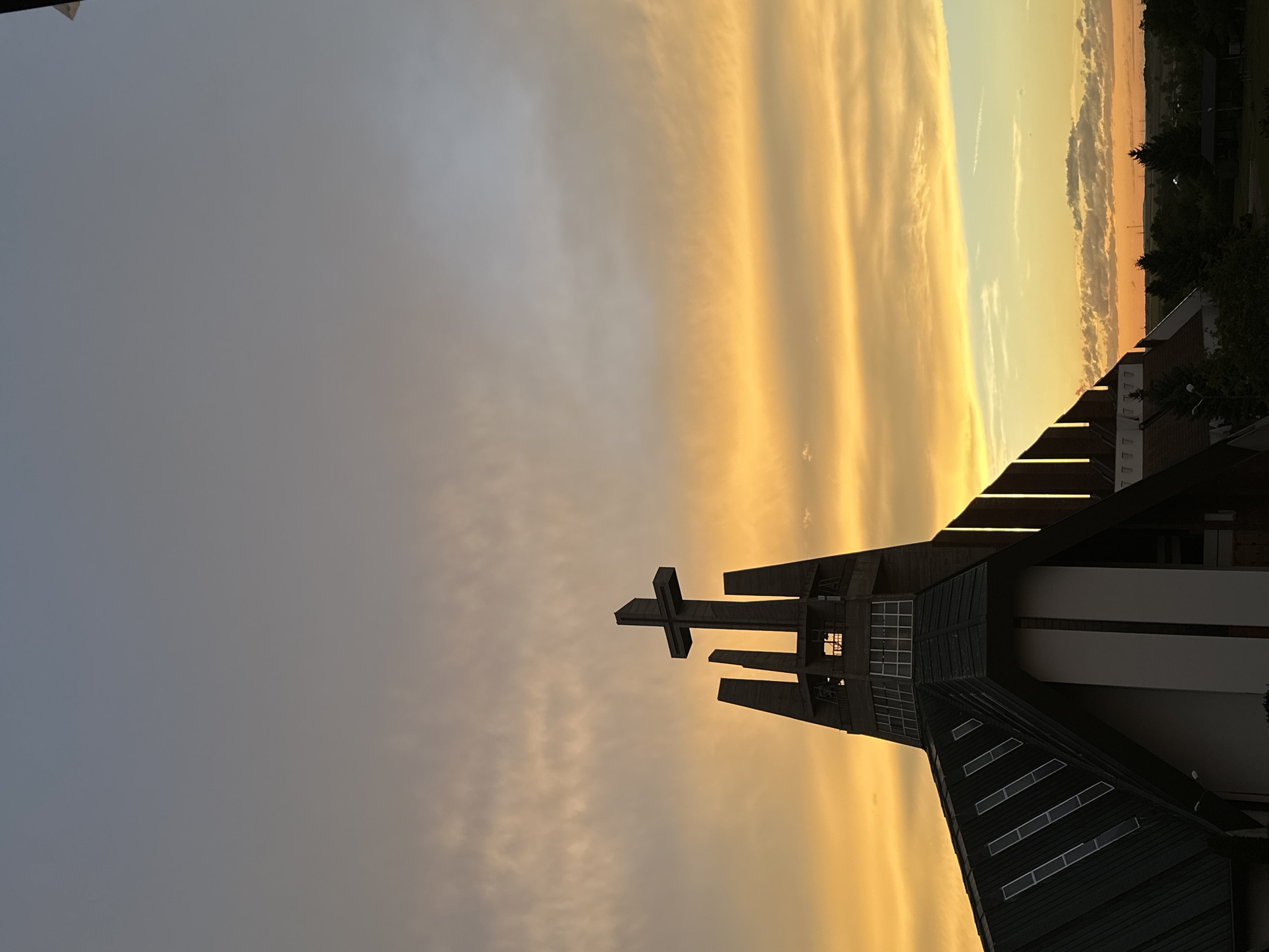 A modern church with a prominent cross on its steeple is silhouetted against a sunset sky. The sky is filled with dramatic, colorful clouds in hues of orange, yellow, and blue. The church features a distinctive architectural design with angular lines and large windows. The surrounding landscape appears to be a mix of trees and buildings, suggesting an urban or suburban setting. The image captures the serene beauty of the sunset over the church. This is the Church of the Holy Trinity in Świdnica, Poland.