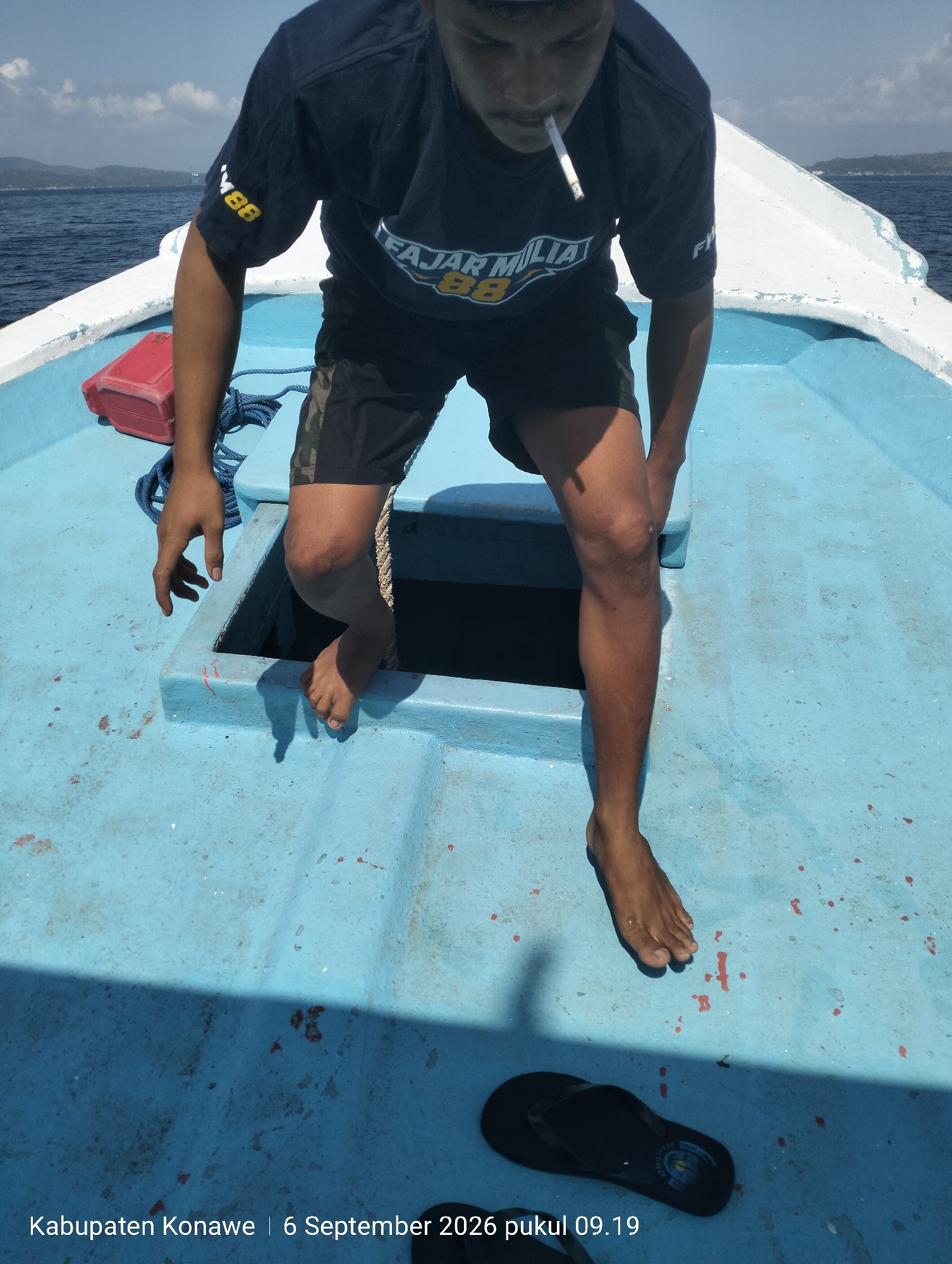 The image is a medium shot from a high angle of a person on a boat. The person is wearing a dark t-shirt with a yellow emblem and black shorts. They are smoking a cigarette and have one foot inside a blue opening in the boat. The boat is on the water, with a shoreline visible in the distance. The text at the bottom of the image indicates the location as "Kabupaten Konawe | 6 September 2026 pukul 09.19".