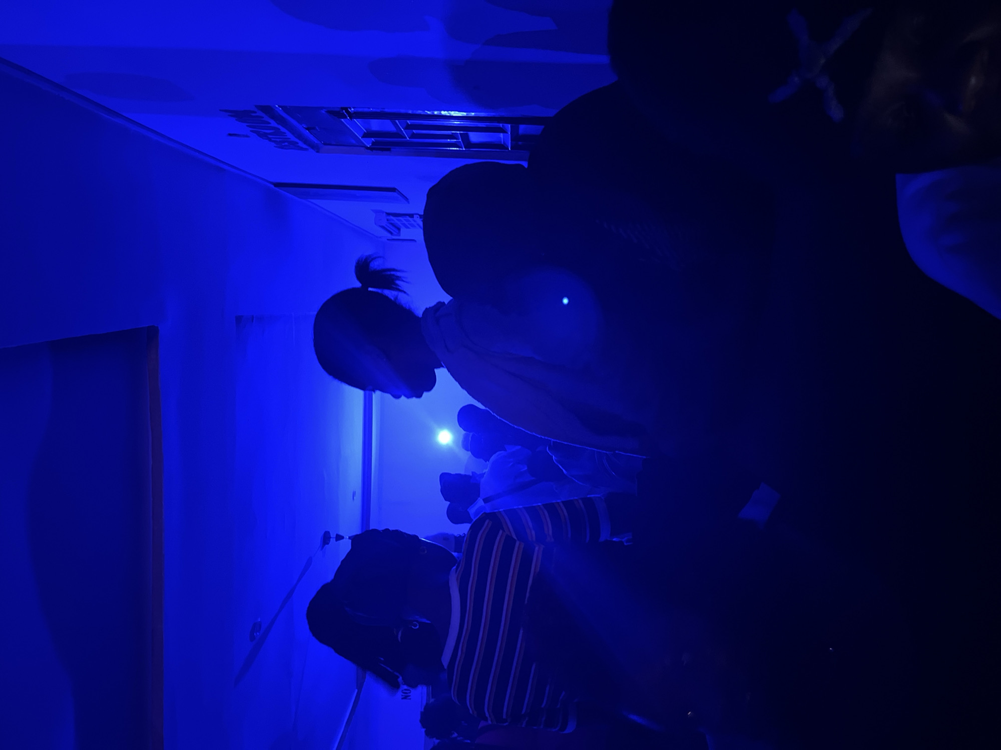 The image depicts a dimly lit indoor scene dominated by deep blue lighting. Silhouetted figures are present, some with distinct hairstyles like ponytails. One individual in the foreground is wearing a horizontally striped shirt with orange and white bands. Another person is partially visible in the center, facing to the right, with their hair pulled back in a ponytail. In the background, there are more indistinct figures and a sign on the wall that reads "RESTROOM". The environment appears to be a public space, possibly a venue or event, with what looks like a window with panes to the right. The overall atmosphere is dark and somewhat obscured by the intense blue lighting and shadows. The location in the city of Durumi, Nigeria, is not explicitly visible or indicated within the image itself.