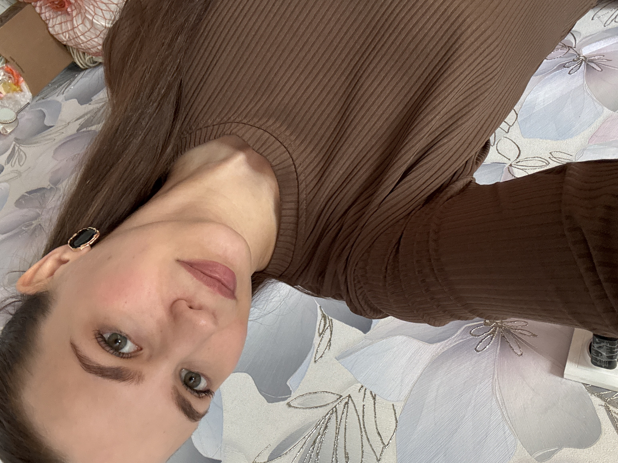 A close-up, rotated portrait shows a young adult woman indoors, likely posing for a selfie. She has long, straight dark brown hair, fair skin, and light green eyes. Her eyebrows are well-defined, and she wears a subtle makeup look with pinkish-brown lipstick. A black oval-shaped earring with a gold-colored rim adorns her left ear. She is dressed in a brown, ribbed long-sleeved top.
The woman is positioned diagonally across the frame, looking directly at the viewer with a neutral to subtly pleasant expression. The background features a patterned surface, possibly wallpaper or bedding, adorned with large, light-colored floral designs outlined in metallic silver or gold. Some small, reflective details are integrated into the floral patterns.
To the upper left of the woman, indistinct items like a light-colored woven object and a small brown box are partially visible. In the lower right corner, a white electrical outlet and a black plug or device are seen. The lighting is soft and consistent with an indoor environment. The setting is indoors in what appears to be a residential or personal space. The location context indicates this image was taken in Nizhnyaya Gar', Russia. No text is visible in the image.