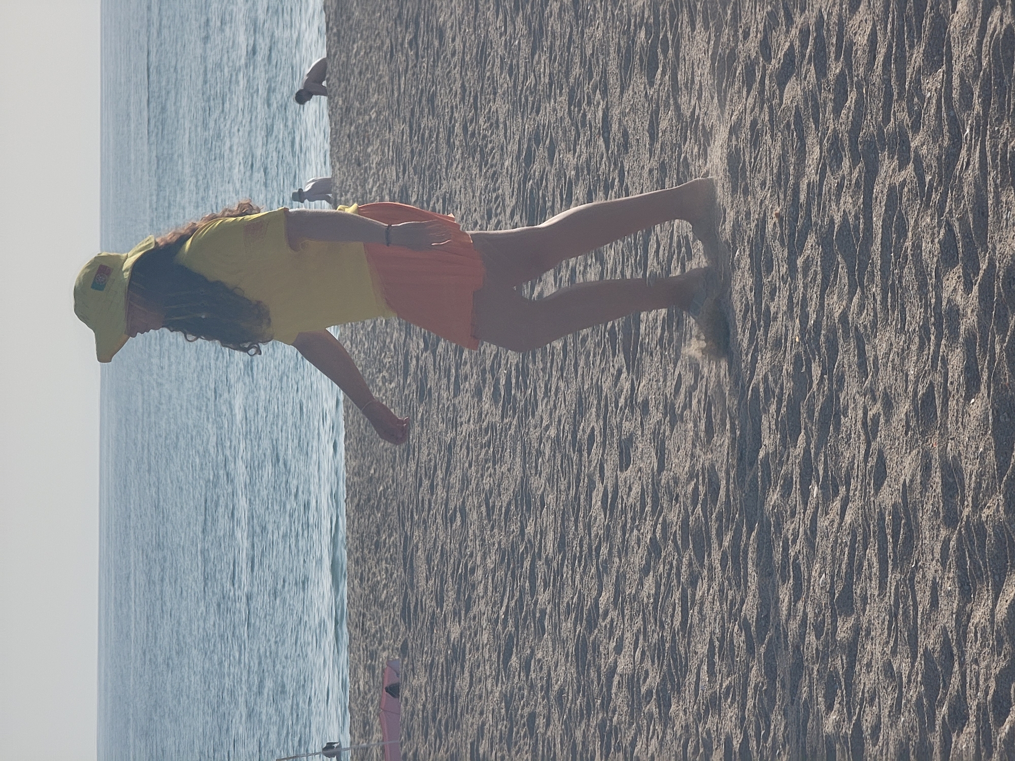 The image depicts a beach scene in Quarteira, Portugal, under a bright, hazy sky. The foreground is dominated by textured sand, with ripples and small debris. A person, possibly a lifeguard given their yellow shirt and cap adorned with a crest, walks across the sand, facing left and away from the viewer. They are wearing an orange skirt and sandals. In the background, the calm, blue ocean stretches to the horizon, meeting a pale sky. A few other individuals are visible further down the beach, appearing as small figures near the water's edge. A red and white object, possibly a float or buoy, and a thin pole are partially visible on the left side of the frame. The overall impression is of a tranquil, sun-drenched day at the coast.