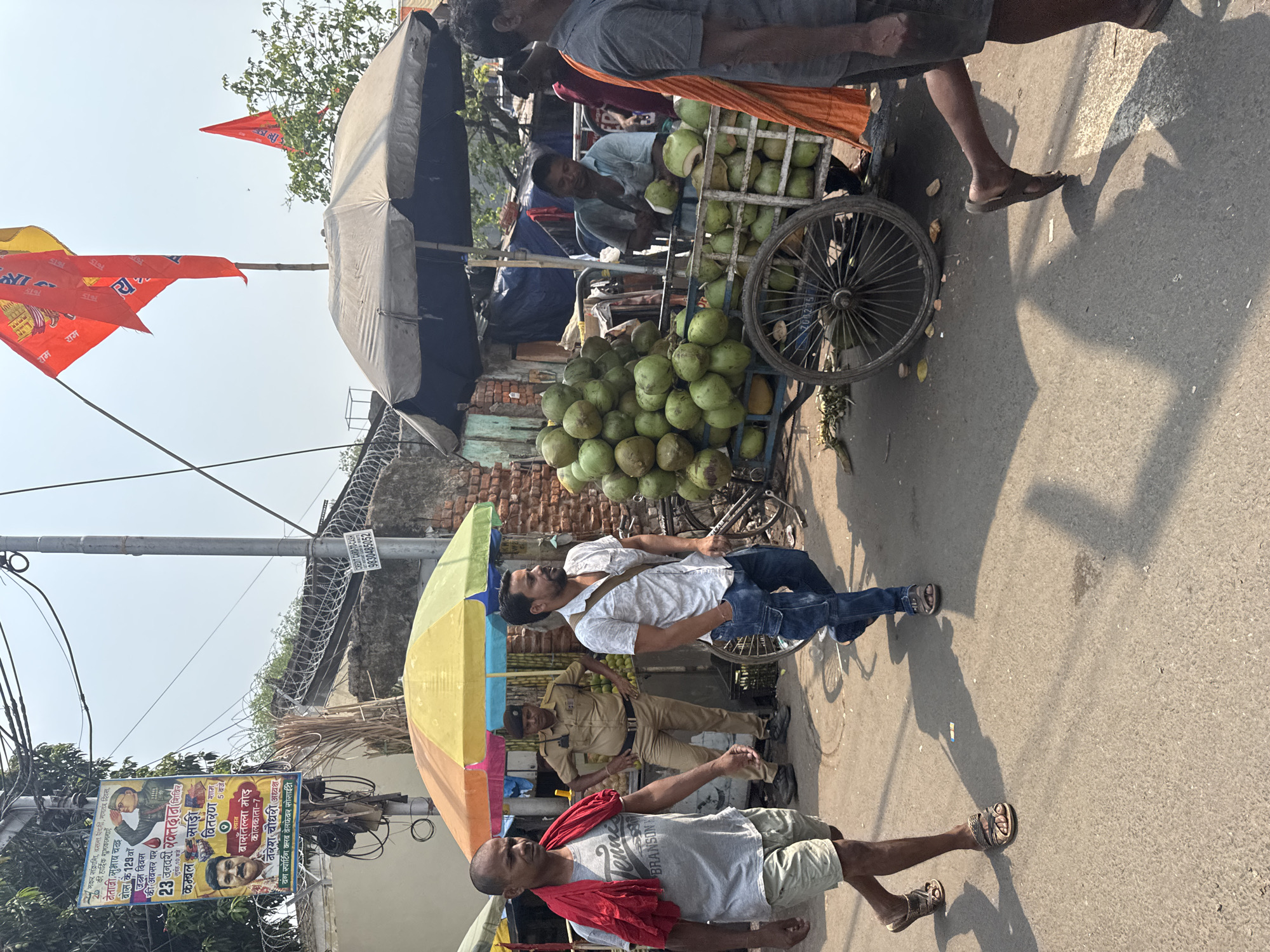 A medium-shot image of a street in Kolkata, India shows several people and structures. A vendor's pushcart laden with green coconuts and covered with a gray tarp is visible. A man wearing a light blue shirt is cutting coconuts at the cart. In front of the cart, a man in a gray shirt and khaki shorts is walking, a red cloth draped around his shoulders. Another man in a white shirt and jeans is sitting on a bicycle. Next to him stands a man in a tan police uniform. An umbrella with multi-colored panels shades them. In the background, a brick wall and metal gate are visible. A utility pole on the left has numerous wires and a poster that reads "मकर संक्रान्ति, वसन्त पंचमी, गणतंत्र दिवस." Several red flags with writing on them flutter in the background. The street is paved and the lighting indicates it is daytime.