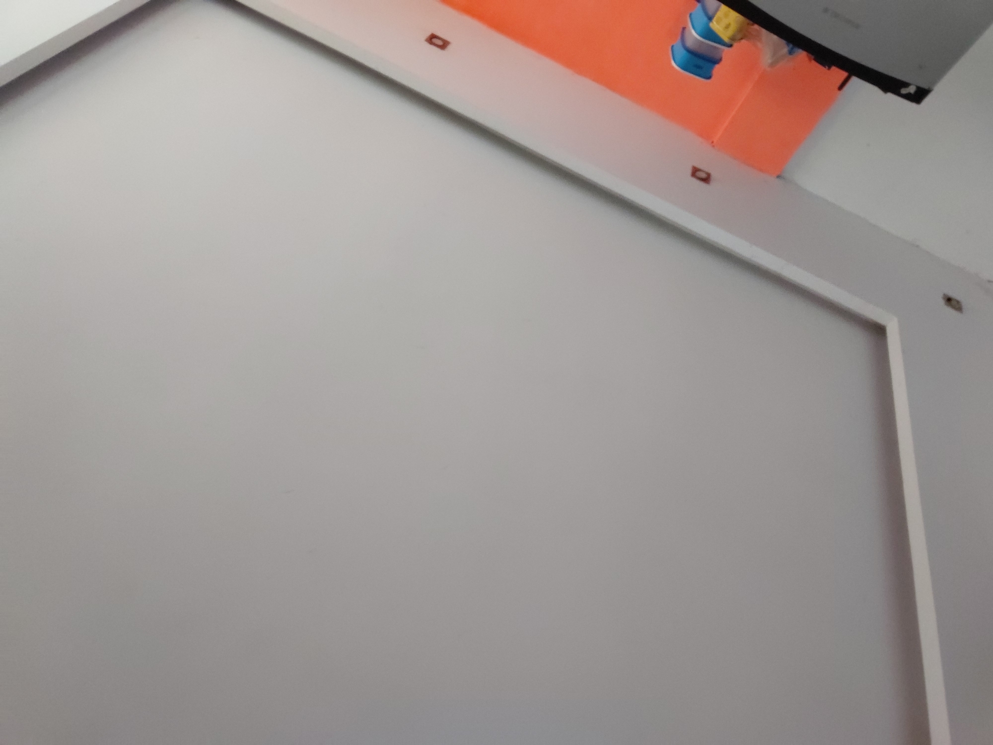 The image captures an indoor scene from an upward and angled perspective, primarily featuring architectural elements. A large, light grey or off-white surface, possibly a wall panel or a section of a cabinet, dominates the foreground and center. This surface is bordered by a distinct white frame or trim, creating a defined rectangular area within the view.
Above this white-framed section, an expansive, bright orange surface extends across the upper part of the image. This orange area could be a ceiling or an upper wall section, adding a vibrant contrast to the neutral tones below. Several small, dark, rectangular details are visible along the lower edge of this orange surface, appearing as mounts, brackets, or decorative elements. One is clearly centered on the left side, another further right, and a third partially seen towards the far right.
In the upper right corner, partially obscured, are a few colorful objects. These appear to be small items, possibly plastic containers or parts of a fixture, displaying shades of blue, yellow, and white. Adjacent to these, a dark, angular object is visible, which could be part of a screen or an electronic device mount.
The lighting throughout the scene is even, suggesting either indoor artificial illumination or diffused natural light. There are no people visible, and no specific activity is taking place. No discernible text is present in the image. The setting is located in Santa Ana, Venezuela.