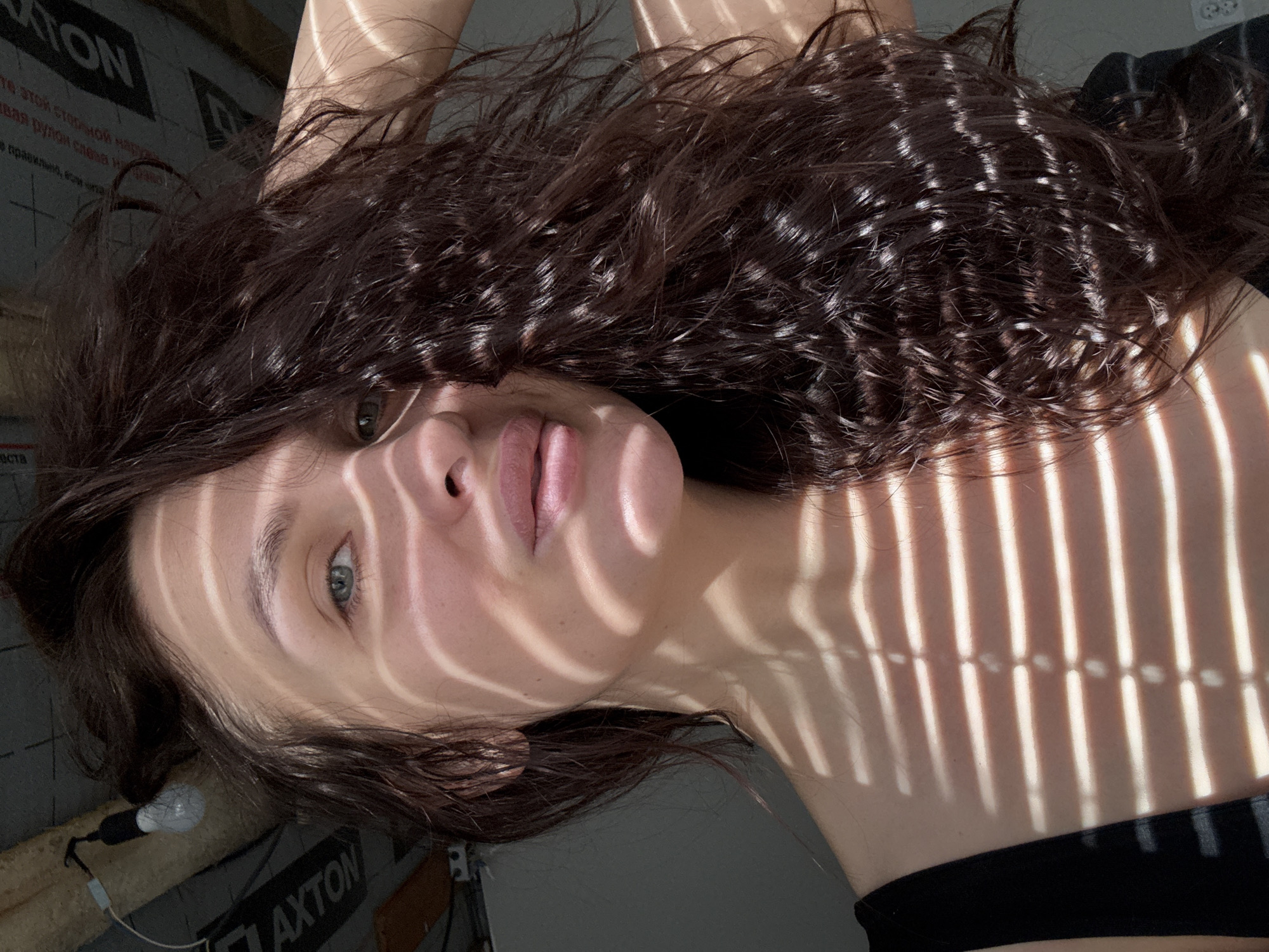A female subject is depicted in a close-up indoor portrait shot in Chaikovsky, Russia. She has dark, wavy hair, and her light skin is illuminated by direct sunlight passing through horizontal blinds, casting prominent parallel striped shadows across her face, neck, and upper chest. Her green eyes are visible through the shadow pattern, and her lips are slightly parted. She is wearing a black top, with a shoulder strap visible on her left side. Her right hand is positioned behind her head, partially obscured by her hair. In the upper left background, an exposed ceiling structure features wooden beams, a dark gray vapor barrier or insulation material with the white capitalized word "AXTON" printed on it, and a single incandescent light bulb hanging by a wire. The background immediately behind the subject is a plain, light-colored wall.