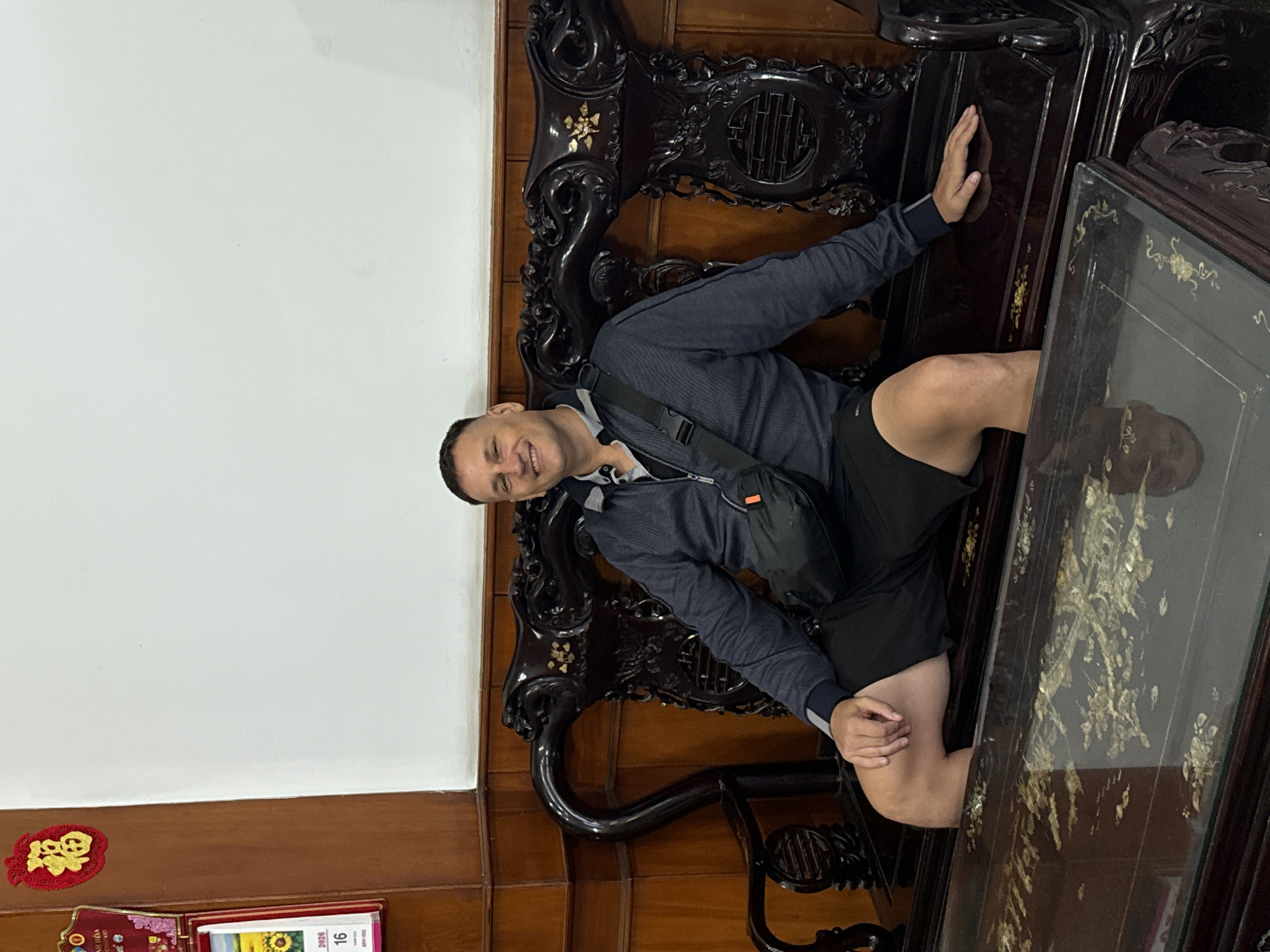 A middle-aged man with short hair is sitting on an ornate, dark wood chair and leaning against a dark wood table with a glass top. The chair and table appear to be antique or traditional furniture. The man is wearing a dark blue jacket, a light-colored shirt underneath, and black shorts. He has a black bag slung across his body. The background includes a white wall on the left and wood paneling behind the furniture. A small red and gold decoration is visible on the lower left side of the image, along with what appears to be a calendar with the number 16. The location is in Tay Nha Trang Ward, Vietnam.