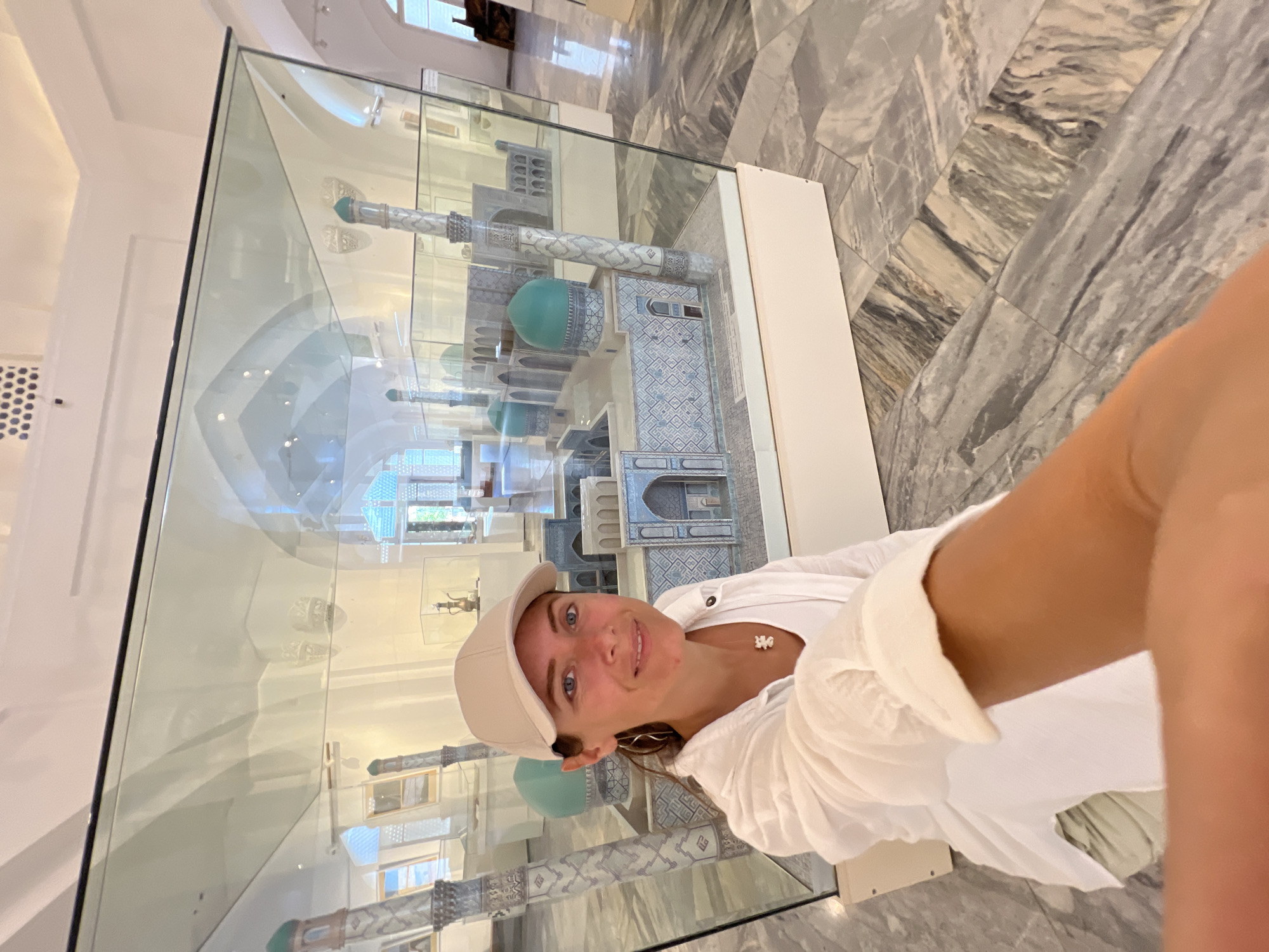 A woman in a white shirt and beige baseball cap takes a selfie in front of a glass display case. Inside the case is a detailed architectural model of a mosque with blue domes and intricate tile work, and tall minarets. The floor is made of polished grey and white marble tiles laid in a herringbone pattern. The background shows more display cases and architectural elements, suggesting an indoor museum setting. The scene appears to be the Gur-e-Amir Mausoleum in Samarkand, Uzbekistan, a historical site known for its stunning architecture.