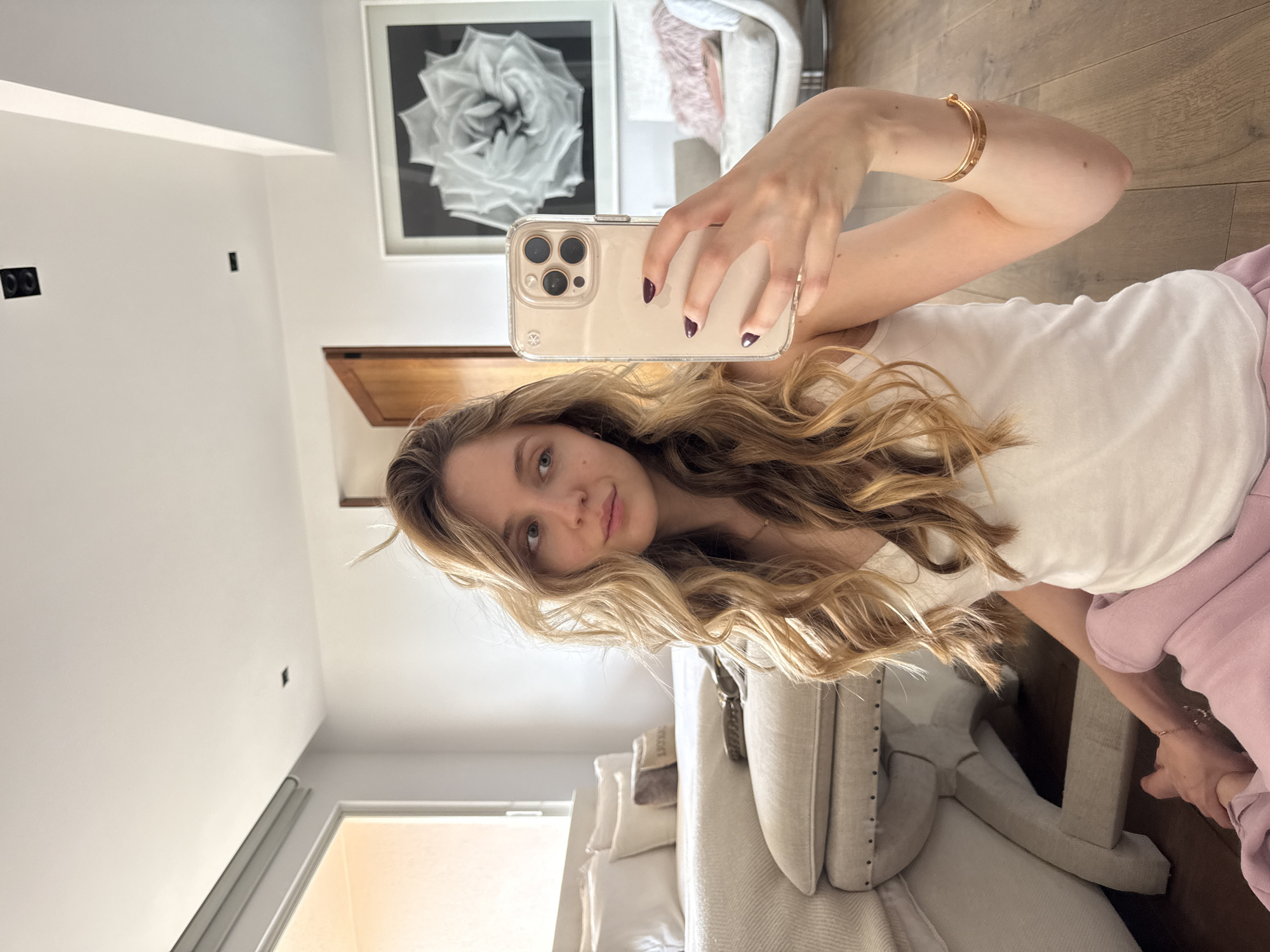 A young woman is depicted taking a selfie while reclining indoors, looking directly at the camera with a relaxed expression and a slight smile. She has long, wavy blonde hair and is wearing a white top and pink bottoms. Her nails are painted a dark plum or burgundy color, and a gold-toned bracelet adorns her right wrist. She holds an iPhone in a clear case, showcasing its multi-lens camera array.
The setting appears to be a modern and well-lit bedroom or living area. The walls are a clean white, complemented by a warm wooden floor. In the background, a prominent black and white framed photograph of a detailed rose is mounted on the wall. Other elements in the room include dark rectangular electrical outlets or switches on a wall, a bed with white pillows and neutral-toned bedding, and a light-colored upholstered armchair or sofa. Natural light filtering through a doorway suggests it is daytime. No visible text is present in the image. The location context is Mexico City, Mexico.