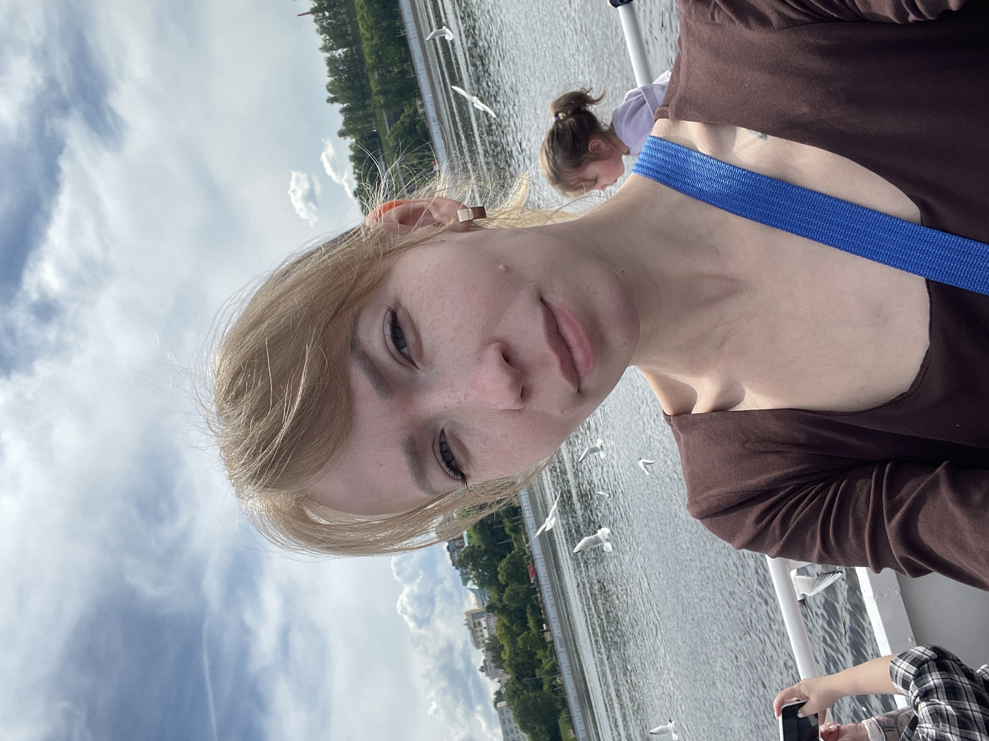 An outdoor portrait shot from a boat captures a young woman in the foreground, facing the camera with a calm expression. She has blonde hair and fair skin with freckles, wearing a brown long-sleeved top and a prominent blue strap crossing her shoulder. A small, square, light-brown earring is visible in her right ear.
In the middle ground, several white seagulls are soaring over a wide body of water, likely a river or large lake. Another person, a child with hair tied in a ponytail, is visible behind the main subject, looking down towards the water. In the bottom right corner, the hands of a third person, wearing a plaid shirt, are seen holding a white smartphone, suggesting they are also taking photos or videos.
The background features a green, tree-lined bank with distant buildings, indicative of a cityscape or town, consistent with a location like Perm, Russia. The sky is partly cloudy, suggesting a daytime setting with fair weather. The scene conveys a relaxed atmosphere during a boat trip or cruise. No visible text is present in the image.