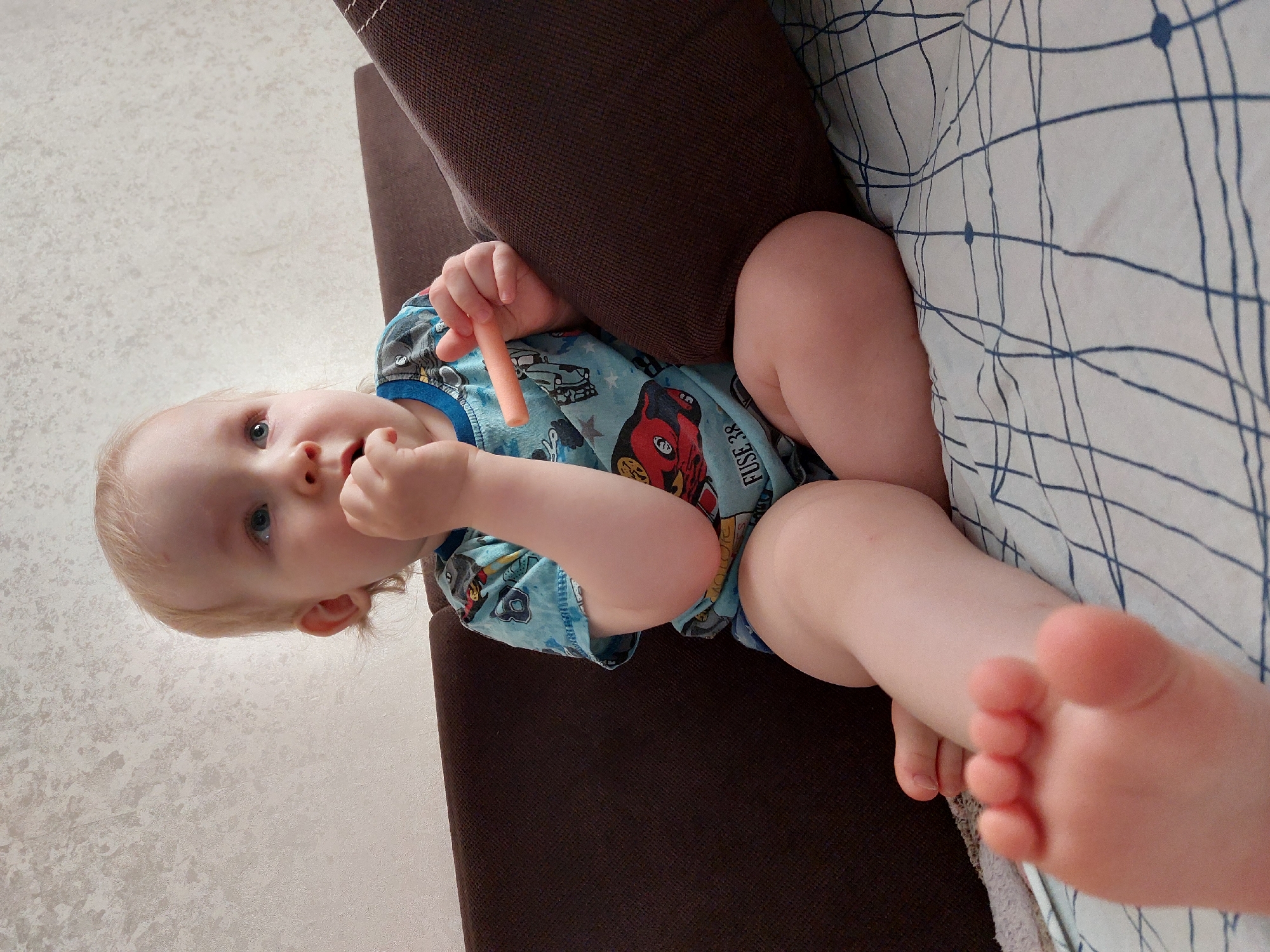 The image, taken in Chaikovsky, Russia, depicts a fair-skinned toddler with light blonde hair and blue eyes, captured from a low-angle perspective. The child is lying on what appears to be a brown sofa or bed, positioned horizontally within the frame, and looking directly at the viewer with a curious or neutral expression.
The toddler is wearing a light blue short-sleeved top adorned with colorful cartoon car illustrations, including a prominent red car design. Partially visible text on the shirt reads "FUSE 38". In one hand, the child holds a small, orange, cylindrical object, possibly a snack stick or toy, near their mouth.
The setting is an indoor space, suggested by the brown textured cushion or armrest, a white sheet with a dark blue linear and circular pattern, and a light beige, textured wall in the background. The child's bare feet are visible in the foreground, with the toes slightly out of focus. The overall impression is one of a calm, everyday moment.