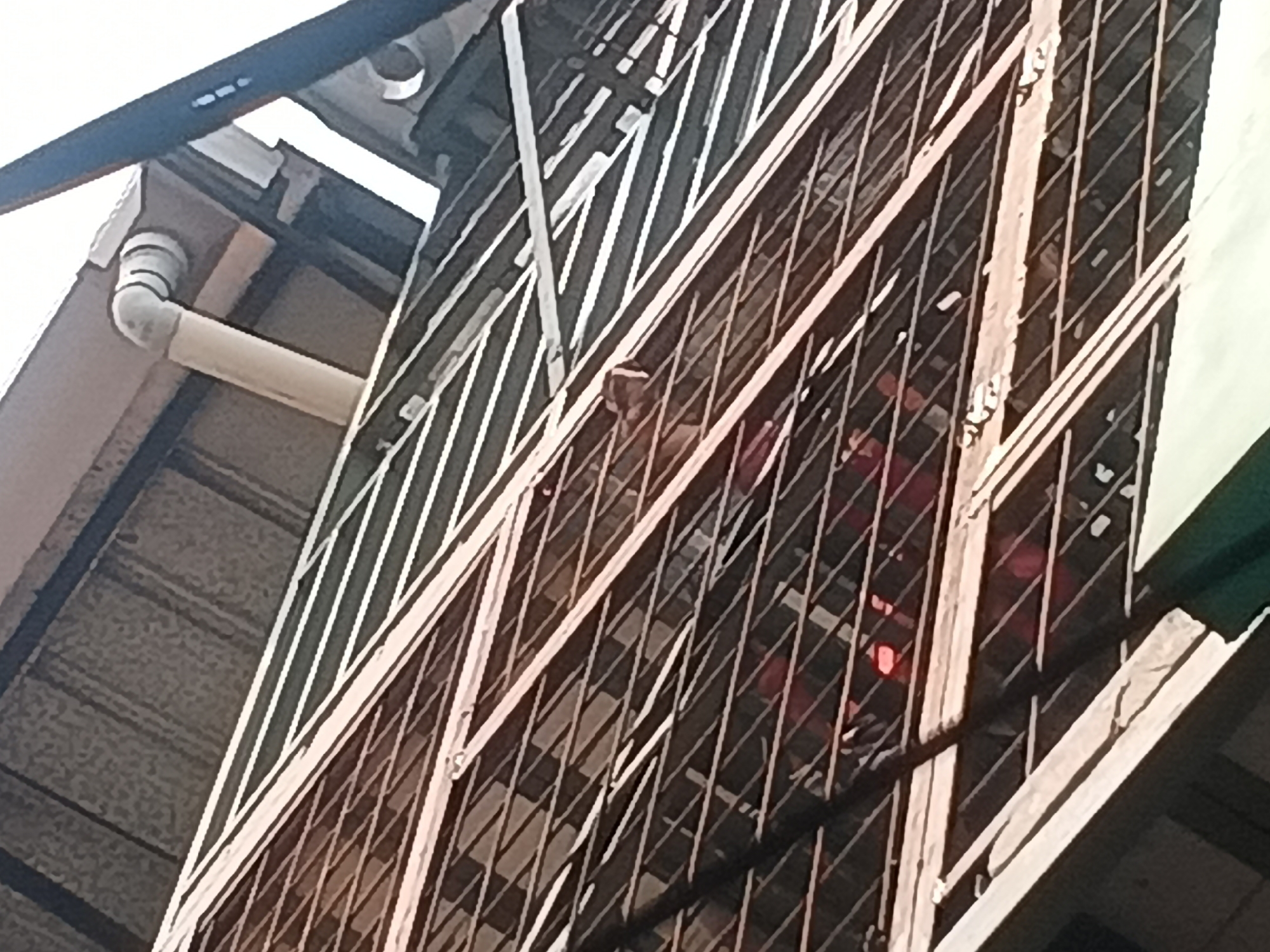 The image captures a close-up view of a metal grillwork structure, possibly part of a building's exterior or a security feature, seen from below. A white downspout pipe is visible on the left side of the frame, curving downwards. Behind the grillwork, there are glimpses of what appear to be people or figures, with some areas showing red or pink hues, suggesting clothing or other colored objects. The overall lighting suggests it is daytime, with some shadows cast by the grill. The scene is likely outdoors, given the angle and the presence of architectural elements. The image does not contain sufficient detail to determine its specific location within West Jakarta, Indonesia.
