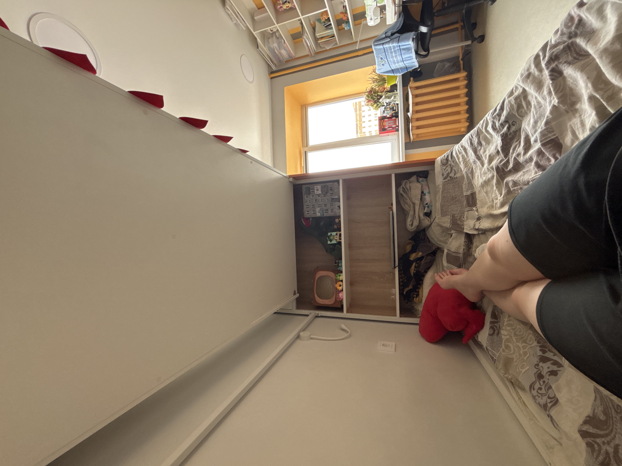 The image shows a room in a building in Perm, Russia. The room contains a bed with a patterned bedspread. A person wearing black shorts is sitting on the bed, their legs visible. Next to the bed is a built-in cabinet. A window is visible in the background, above the cabinet. A shelf filled with various items hangs above the window.