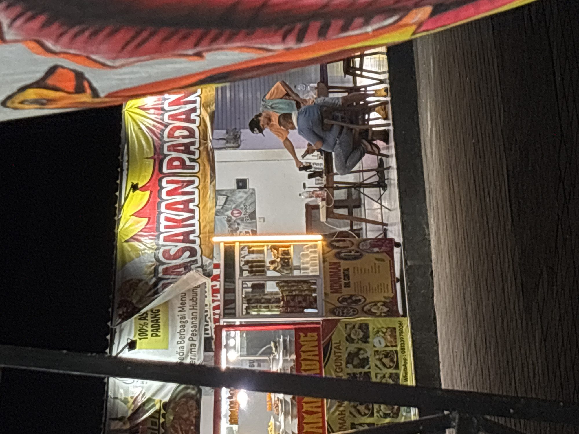 The image depicts an outdoor scene at night, likely in Kuta Selatan, Indonesia, featuring a food stall operation. A banner with "MASAKAN PADANG" and "100% ASLI PADANG" is prominently displayed, indicating a Padang cuisine restaurant. Two men are visible interacting near a table set up with food and drinks. One man, wearing an orange t-shirt and shorts, is standing and gesturing while holding a phone. The other man, seated and wearing a blue shirt and jeans, is looking at his own phone. The food stall has multiple display cases filled with food items and drinks. A bright yellow vertical light is also visible on the right side of the stall. The ground appears to be paved with dark, textured material, possibly asphalt or concrete. A portion of a colorful, large flag or banner with red and orange elements is present on the far right edge of the frame, obscuring part of the scene.