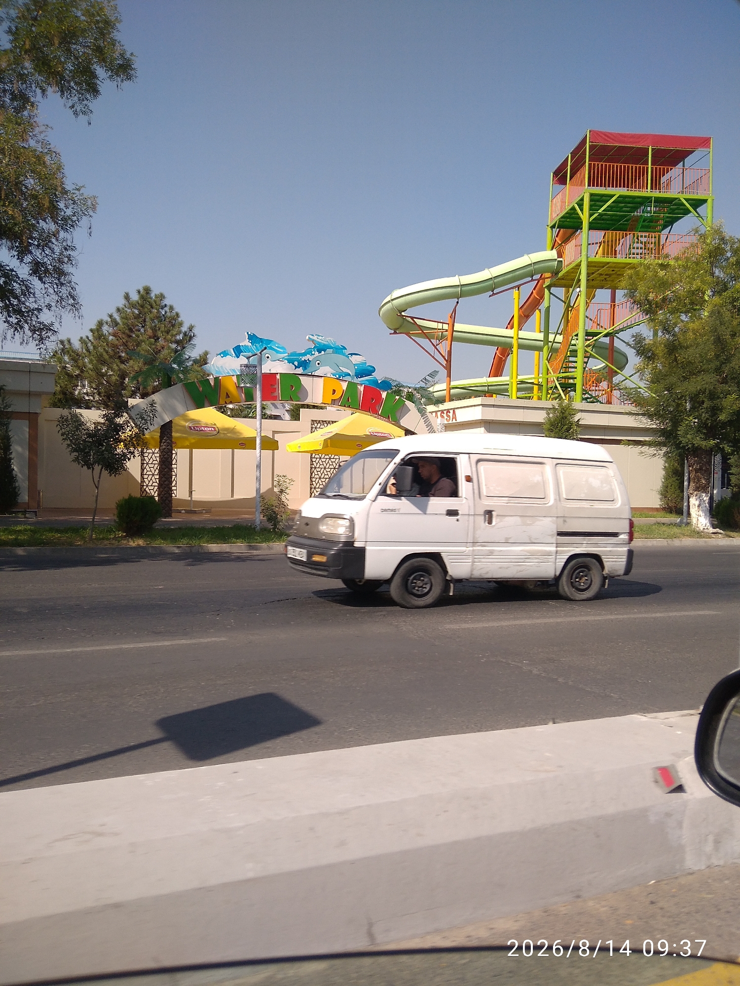 In the city of Tashkent, Uzbekistan, on August 14, 2026, at 9:37 AM, a white van is traveling on a road. The van has tinted windows and at least one person is visible inside. In the background, there is a water park with large, colorful water slides. The park has a sign with the words "WATER PARK" and images of dolphins. Yellow umbrellas with the Lipton logo are also visible. The sky is clear and blue, and trees line the street. The image also shows a shadow of a pole on the road, and a section of a concrete curb in the foreground.