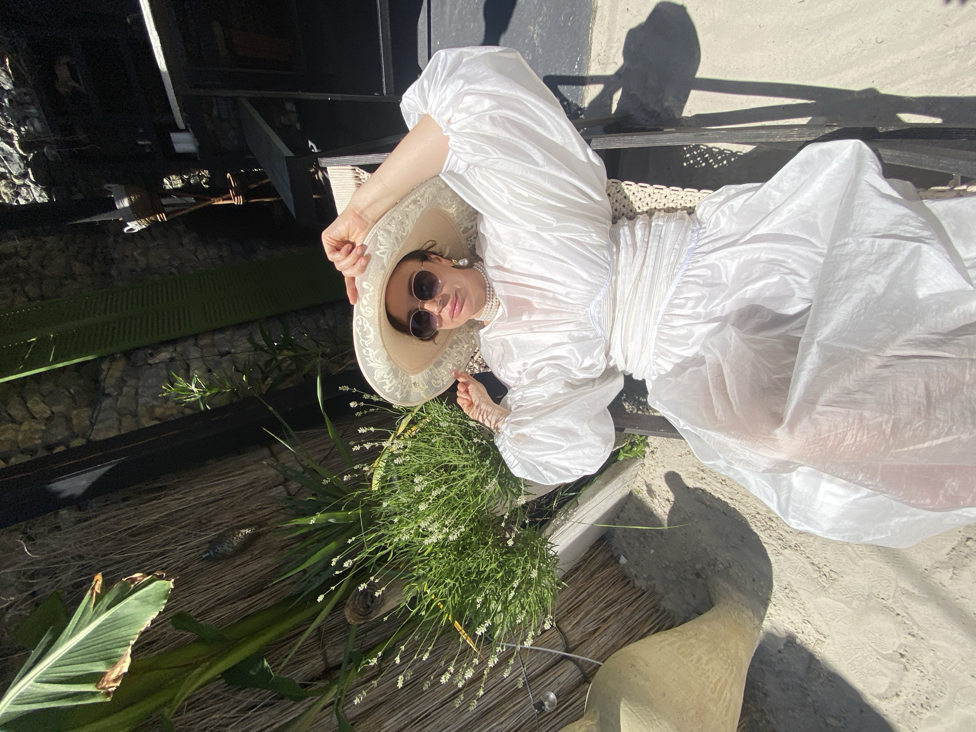 The image shows a woman wearing a large, ornate white hat and a voluminous white dress, sitting in a chair outdoors. She is wearing sunglasses and a pearl necklace. The background includes a thatched wall, greenery, and a stone wall, suggesting an outdoor seating area. The ground is sandy. This scene appears to be located in Sochi, Russia, specifically within the "Riviera" Park, known for its lush vegetation and diverse recreational spaces. The lighting indicates it is a sunny day, casting shadows on the sand.