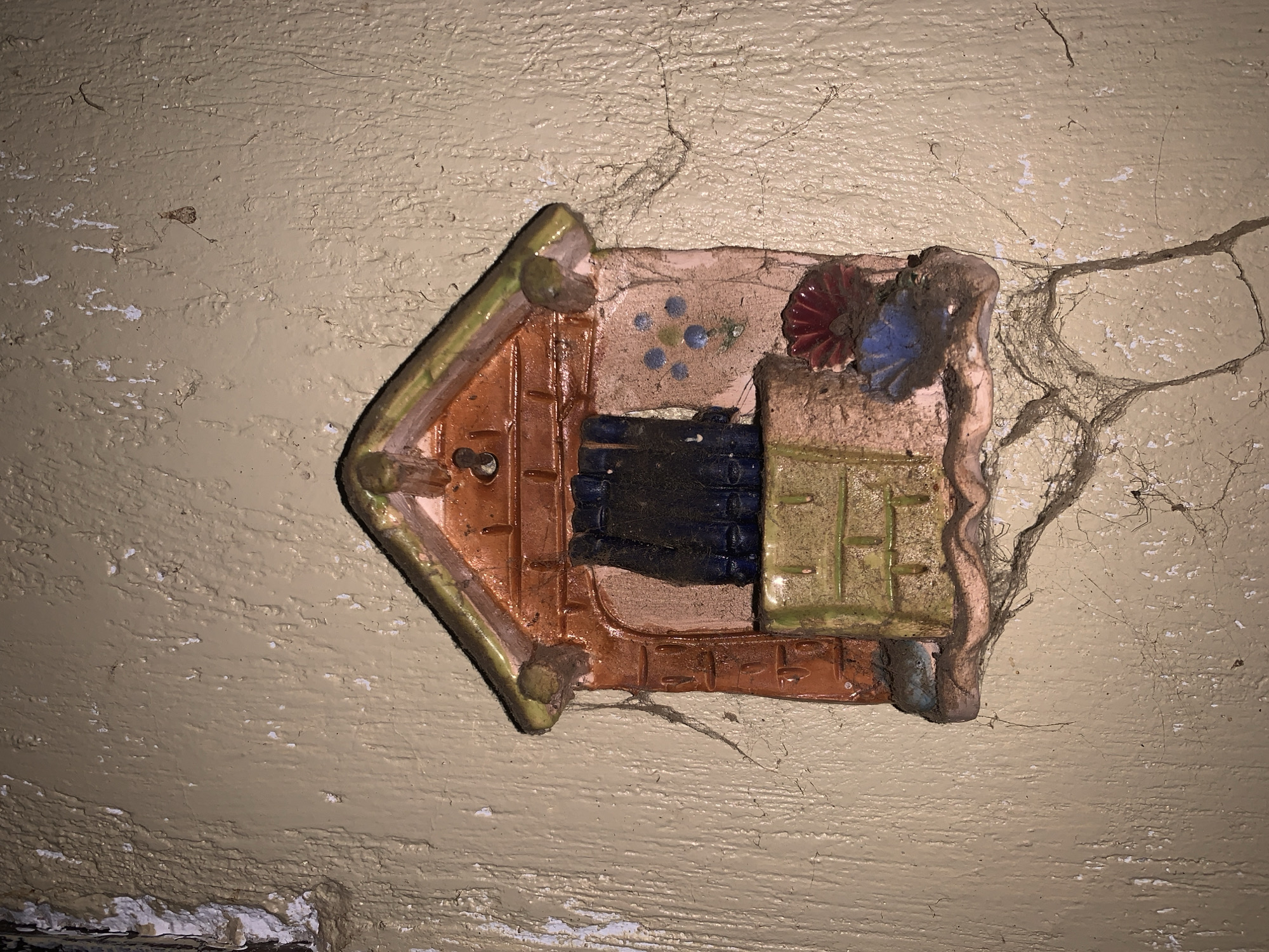 This media file captures a close-up view of a weathered ceramic wall decoration mounted on a textured, light beige wall in Santa Ana, Venezuela. The decoration is arrow-shaped, pointing towards the left, and designed in a relief style to resemble a miniature house or building facade. It features a green and brown pointed section on the left, forming what appears to be a roof. The main body of the decoration is a reddish-brown, mimicking brickwork or wooden panels, with a prominent dark blue section likely representing a window or door. Towards the right, there's a light brown area with small blue circular patterns, possibly depicting flowers, and another section with green horizontal lines. A small, intricate floral design in red and blue sits on the far right of the decoration.
The entire surface of the decoration and the surrounding wall is heavily covered in dust, dirt, and numerous cobwebs, indicating a long period of neglect. The wall itself shows significant signs of age and wear, with extensive peeling white paint and noticeable cracks spreading across its surface. Within the visible cobwebs, particularly in the upper right area and in the provided cropped sections, several small spiders can be faintly discerned. The lighting is dim and appears artificial, highlighting the textures of the wall and the dust accumulation. There is no discernible activity or event taking place, and no visible text. The scene conveys an atmosphere of antiquity and disuse.