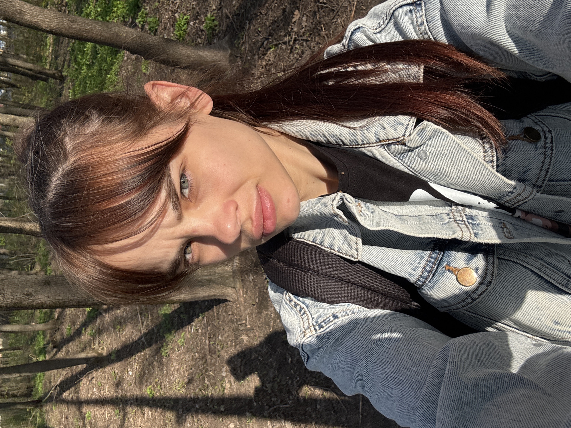 A close-up, slightly tilted selfie captures a young woman outdoors in what appears to be a park or forest in Rostov-on-Don, Russia. She is looking directly at the camera with a neutral expression, her lips slightly parted.
The woman has long, dark brown hair with lighter, reddish-brown highlights visible around her face and along her part, with bangs framing her forehead. Her eyes are light-colored, appearing to be blue or grey-green. She wears a light-wash denim jacket over a dark t-shirt or top, and a black backpack strap is visible over her right shoulder.
The background shows a natural setting with tree trunks and branches, some with hints of green foliage, suggesting spring or early summer. The ground is covered with dried leaves and sparse vegetation. Strong, direct sunlight illuminates the scene, casting distinct shadows on the ground and across parts of her face and jacket, indicating a sunny day, likely during late morning or afternoon. No text is visible in the image.