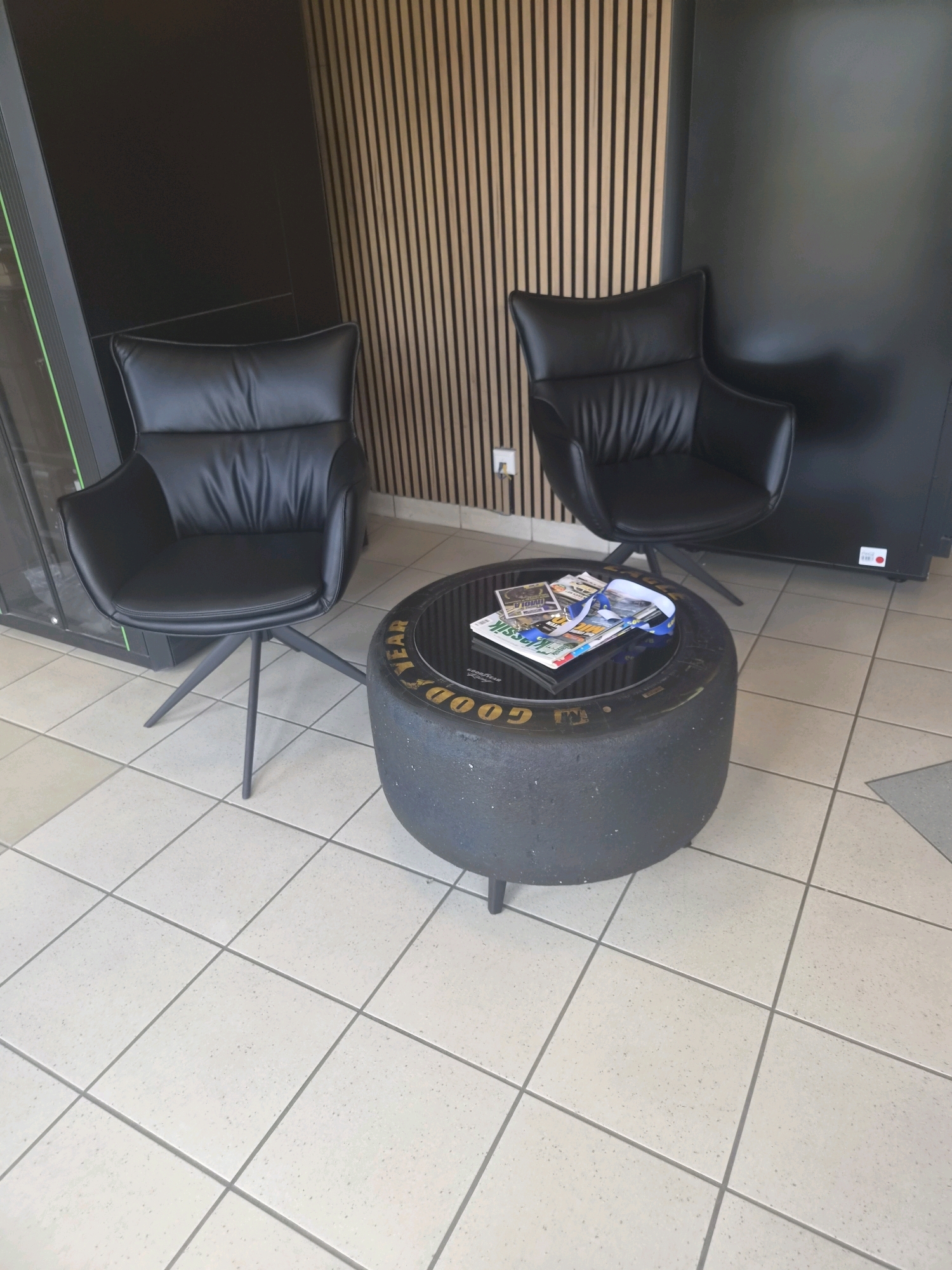 The image depicts a waiting area with two black leather armchairs and a round coffee table designed to resemble a tire. The coffee table has a glass top and magazines are placed on it. The walls behind the chairs are a combination of dark paneling and vertical wooden slats. The floor is tiled with light-colored square tiles. The scene appears to be indoors, likely a commercial or waiting area. The overall aesthetic suggests a modern and perhaps automotive-themed design. The location is identified as Sanem, Luxembourg.