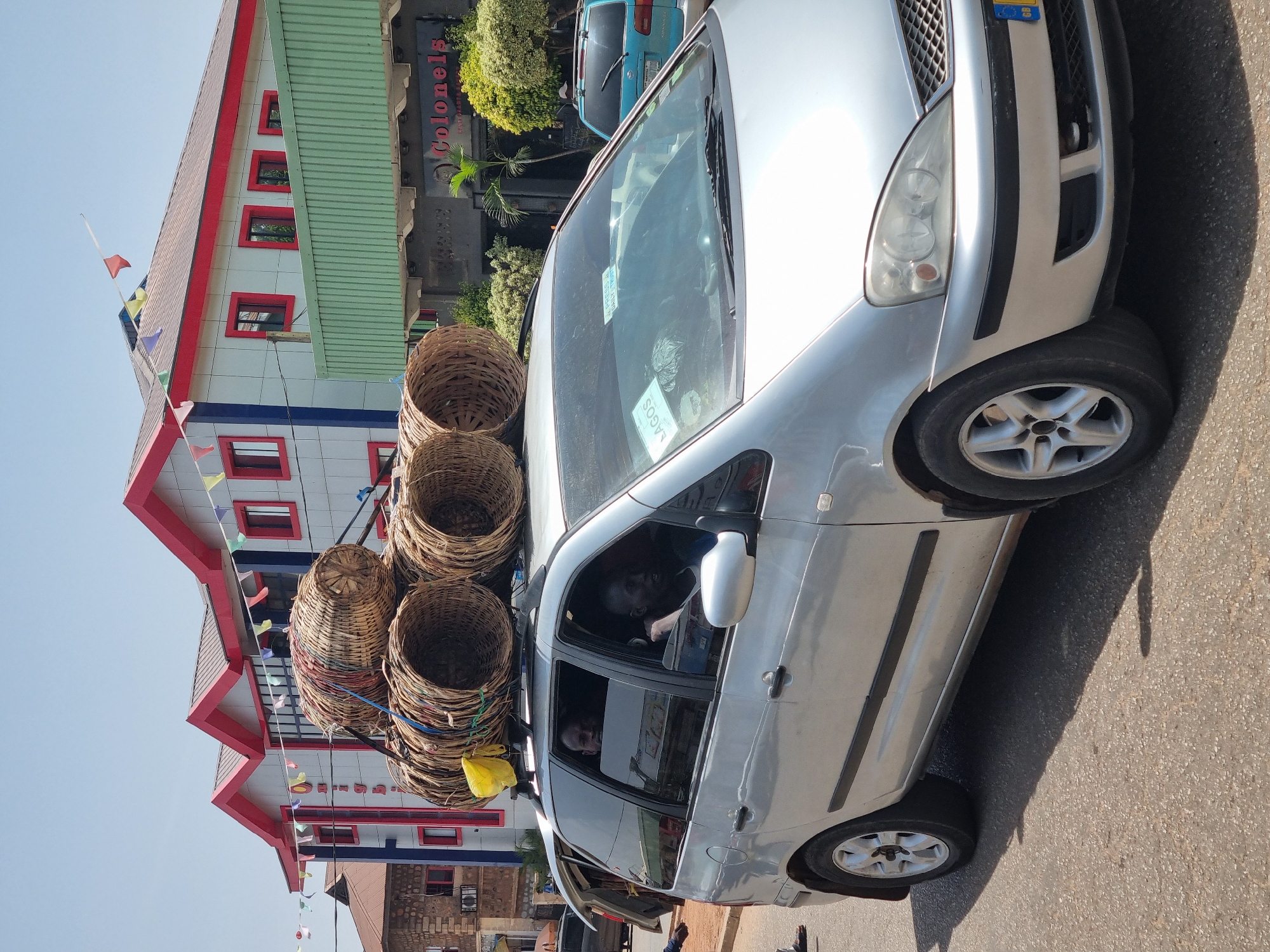 A silver minivan is overloaded with large woven baskets stacked on its roof, secured with ropes, traveling on a street in Jos, Nigeria. The driver and passenger are visible inside the vehicle. A multi-story building with a green roof and red-framed windows is partially visible in the background, along with signage that includes the word "Colonels." The car has a license plate and stickers on the windshield. The street is paved, and a partial view of another vehicle is present. The sky is clear and bright.