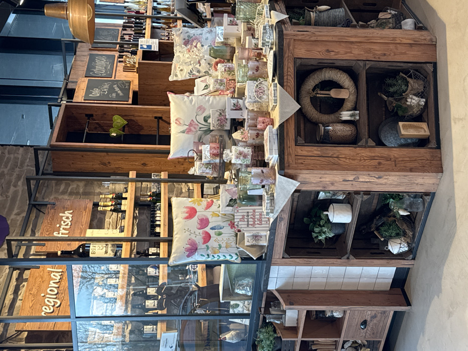 This image displays the interior of a retail establishment in Altenheim, Germany, characterized by a blend of rustic and modern design. The central focus is a large, rectangular wooden display table, constructed from stacked crate-like structures, positioned on a light-colored, smooth floor.
The top surface of this table is covered with a light fabric runner and densely arranged with merchandise. Prominent items include three square throw pillows featuring vibrant floral patterns (tulips and mixed wildflowers), numerous glass candle holders, votives, and small vases, many adorned with floral designs. Various small packaged gift items and a vertical sign with the German text "Du musst dein Anderes Leben" are also present.
In the background, against a stone wall, are dark metal shelving units with wooden shelves. To the left, a glass partition reflects outdoor elements and partially obscures more shelves holding dark-bottled products, with a small white ceramic chicken figurine visible behind it. To the right, wooden signs reading "regional" and "frisch" (fresh) are affixed to the shelving units, which display a variety of bottled goods including a large bottle, champagne, and numerous smaller bottles. Further right, more dark wooden shelves feature chalkboards labeled "Küche" (Kitchen), "Schönes" (Beautiful), and "Getränke" (Drinks), showcasing additional bottled items. A copper-toned pendant lamp hangs from the ceiling in the upper right.
The lower sections of the central display table, configured as open shelves, contain decorative arrangements. These include potted green plants, a white lidded container, a large glass jar filled with granular material, a natural fiber wreath with a small wooden utensil, two white pillar candles, and various baskets holding greenery and a wooden brush. No individuals are present, and no actions or interactions are occurring.