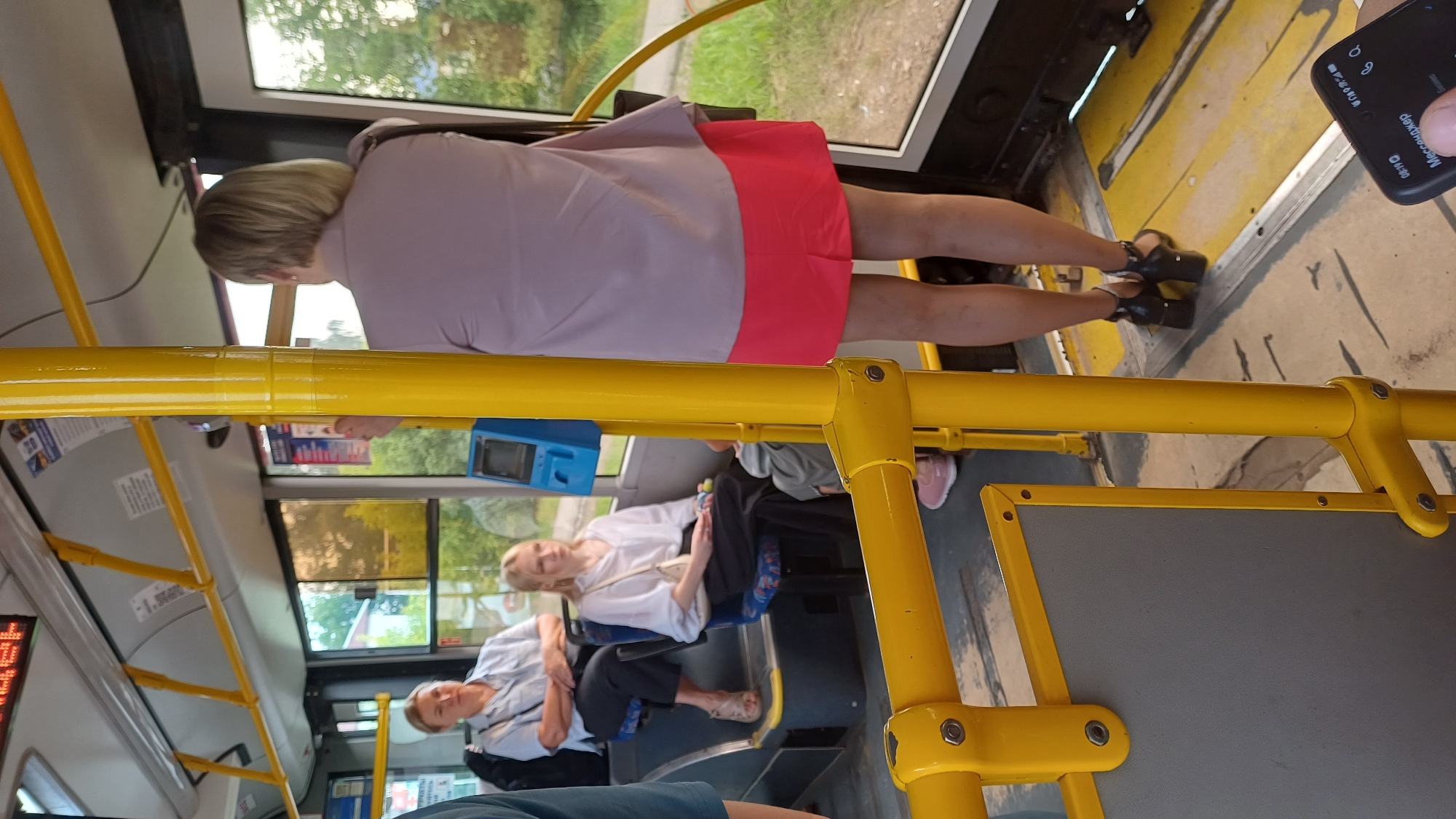 The image captures the interior of a public bus, likely in Izhevsk, Russia, given the context. In the foreground, a yellow pole, a common fixture in public transport, is prominently visible. Behind it, to the left, a woman with blonde hair sits on a blue seat, wearing a light-colored shirt and dark pants. To her left, an older woman in a light blue shirt and dark pants is also seated. In the center of the frame, a young woman with blonde hair is seated, wearing a white blouse and black pants, holding a small object in her hand. To the right, a woman with blonde hair, seen from the back, is standing. She wears a light-colored blouse and a vibrant pink skirt, with black heeled sandals. The bus interior shows yellow handrails and blue seats, with windows revealing a green, tree-lined exterior, suggesting a daytime scene. A blue ticket validator is mounted on the wall. In the lower right corner, a hand holds a smartphone displaying what appears to be a messaging application.
