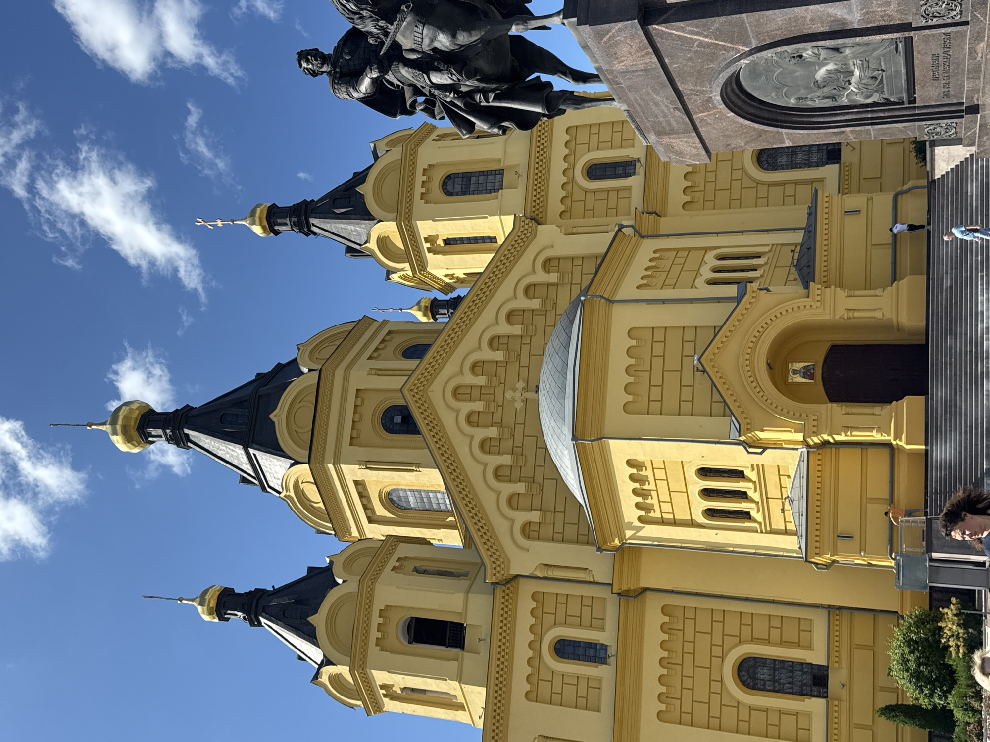 This image shows the exterior of the Alexander Nevsky Cathedral in Nizhny Novgorod, Russia, on a sunny day. The cathedral is a large, yellow Orthodox church with multiple black and gold domes. A bronze equestrian statue of a man in armor is visible in the foreground on the right, with a dark stone pedestal featuring relief carvings. Steps lead up to the cathedral's entrance. Several people are present, including a woman in the lower left foreground looking towards the camera, and a few figures on the steps and near the base of the cathedral. The sky is blue with scattered white clouds. The scene appears to be a tourist or public gathering place.