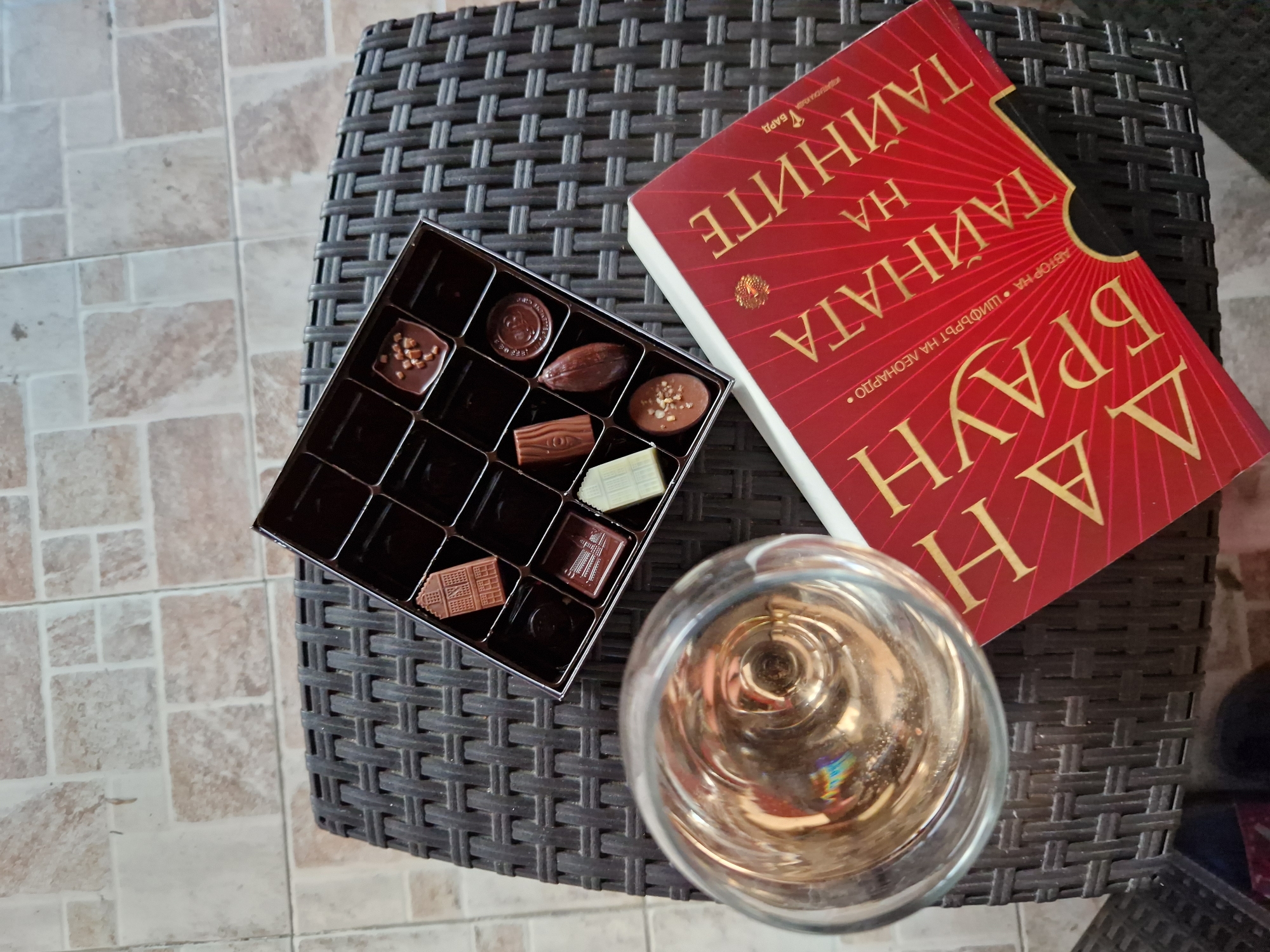 The image shows an overhead view of a glass of champagne, a box of chocolates, and a book with a red cover on a dark wicker table. The book has gold lettering in Cyrillic script. The table is placed on a tiled floor. The scene is indoors, possibly in a cafe or a home. There are no people visible in the image. The overall impression is one of a relaxed and luxurious setting.