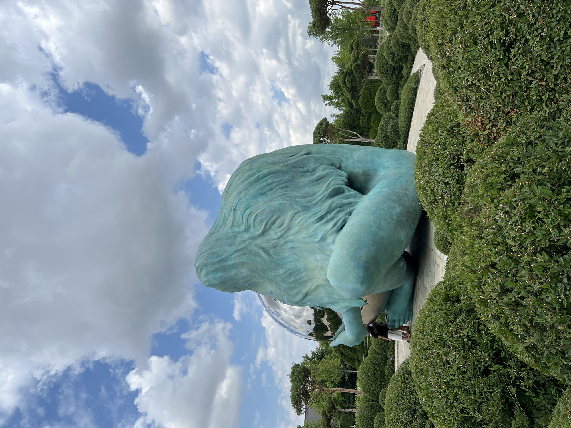 A large, teal-colored sculpture of a seated figure holding a mirrored sphere is the central subject of the image. The sculpture has long, flowing hair and appears to be made of bronze or a similar metal, with a patina giving it the teal hue. Two small figures, appearing to be people, stand at the base of the sculpture, looking up at it. The sculpture is situated in a park-like setting with manicured green bushes and trees. A paved pathway winds through the landscape. In the background, more trees and some red structures, possibly part of a building or another art installation, are visible. The sky above is partly cloudy with patches of blue. This scene is located in the Galitsky Park in Krasnodar, Russia.