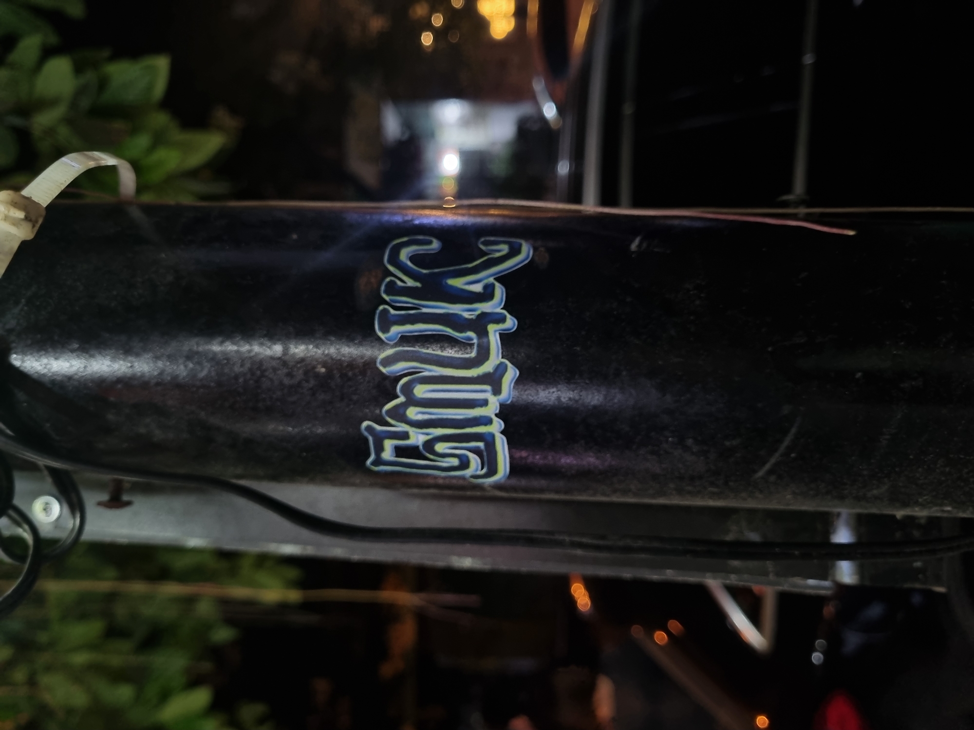 The image captures a close-up, low-light view of a black, textured surface, likely a metal bar or frame, set against a dark, out-of-focus background. The primary subject is a stylized sticker affixed to the center of this surface. The text on the sticker appears to read "HULK," written vertically from top to bottom. The letters are rendered in a distinctive blue color with a bright, light green or yellow outline, giving them a bold, almost three-dimensional appearance.
To the far left of the surface, a white plastic zip tie is visible, partially secured around the black object. Further right and above the sticker, a faint, reddish-brown thread or wire runs horizontally across the surface. The background is heavily blurred, displaying numerous bokeh lights, suggesting an urban or semi-urban outdoor setting at night. Hints of dark foliage are visible on the left side of the background. The surface itself shows some wear, with minor scratches or dust evident. No specific activity is occurring, and the scene is static. The context provided specifies the location as Đà Nẵng, Vietnam.