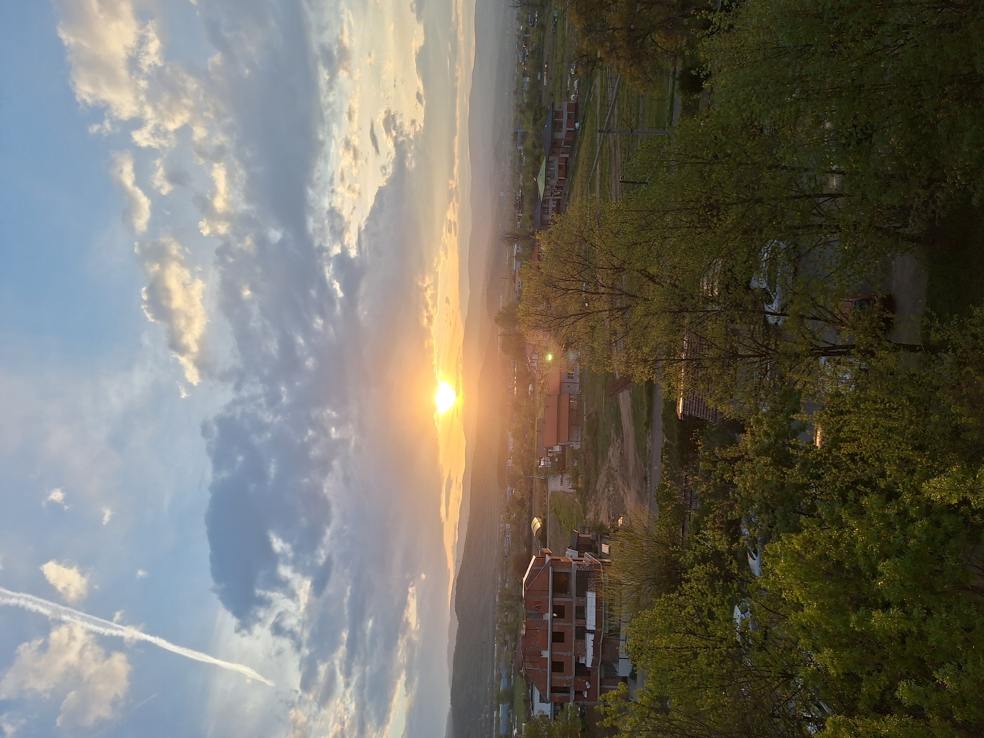 This image captures a vibrant sunset over a semi-urban landscape, likely taken from an elevated vantage point in Blagoevgrad, Bulgaria.
The central subject is the setting sun, a brilliant golden orb low on the horizon, casting a strong, warm glow across the scene. The sky is partially cloudy, with cumulus clouds illuminated in shades of orange, yellow, and white near the sun, transitioning to blue and grey further away. A distinct white contrail from an aircraft stretches across the upper left portion of the sky.
Below the horizon, distant mountains or hills are visible, appearing hazy and silhouetted against the bright sky. The midground features a sprawling town or village with numerous residential buildings scattered across the terrain. Some structures appear to be fully built homes with dark roofs, while others, particularly in the foreground, are red-brick building frames in various stages of construction, indicating ongoing development.
The foreground is dominated by lush green trees with vibrant foliage, obscuring parts of the town below. Their leaves are brightly lit by the low sun, creating a lively contrast with the shadows. Roads or paths wind through the settlement, and some utility poles or power lines are also visible.
The overall scene depicts the peaceful end of a day, with the soft, golden light of sunset painting the clouds and illuminating the developing community. No discernible text is visible in the image.