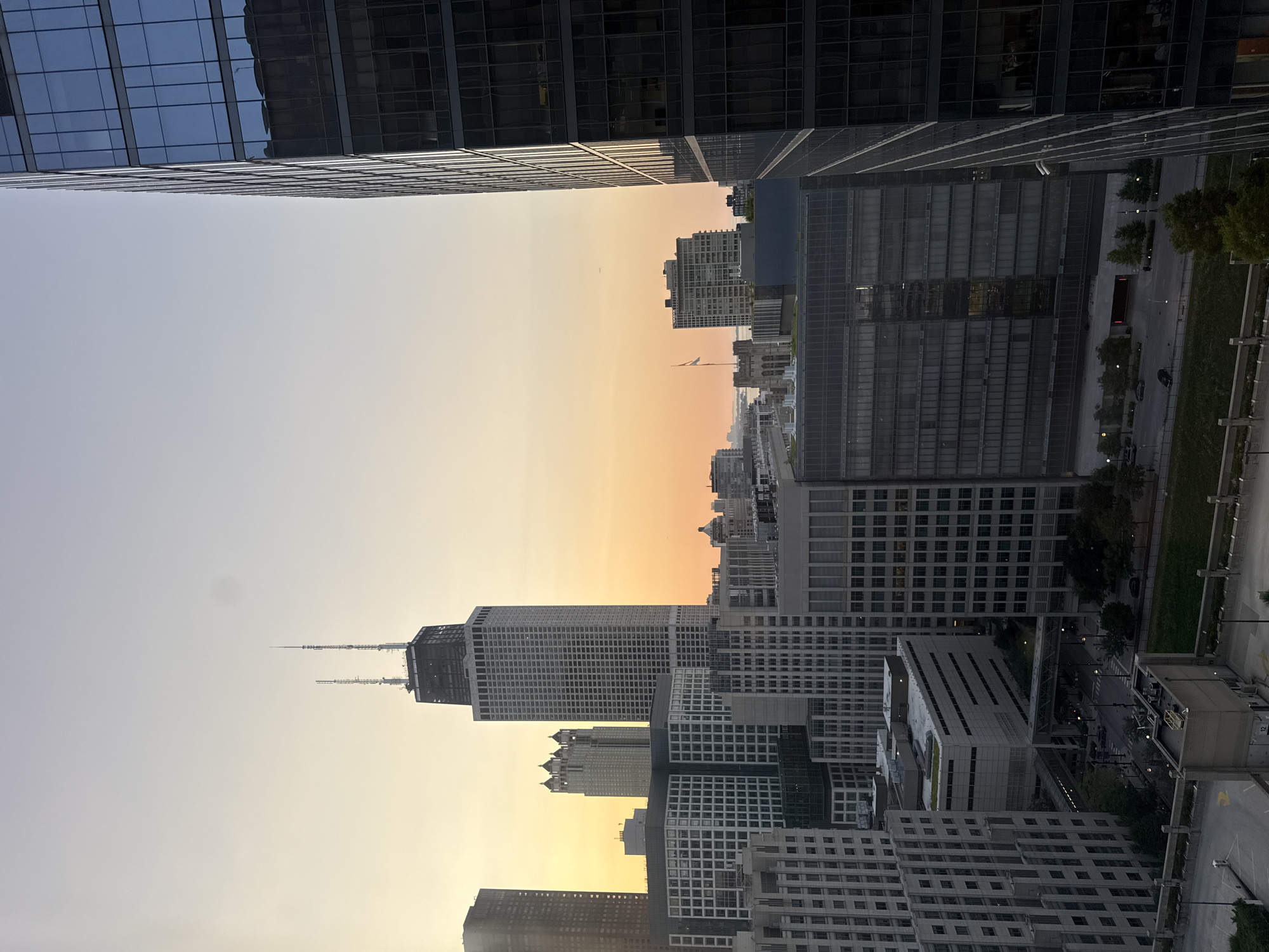 This is an exterior, eye-level, medium shot of the Chicago skyline during a clear, hazy sunset. The image is dominated by various skyscrapers. In the center of the frame, the iconic John Hancock Center stands tall, its distinctive silhouette featuring twin antennas at its apex. To its left, other buildings of varying heights and architectural styles are visible, including a cluster of buildings with ornate tops and a long, dark-colored skyscraper.
To the right, a modern, glass-paneled skyscraper occupies a significant portion of the right side of the image. Its reflective facade mirrors the sky and surrounding buildings. In the foreground, other mid-rise buildings with grid-like window patterns are visible, suggesting a dense urban environment. The sky transitions from a pale blue at the top to a warm orange and yellow hue near the horizon, indicating the setting sun. A few distant buildings and what appears to be a body of water are visible in the background. No people are discernible in the image. The overall scene captures a typical urban panorama of Chicago at dusk, characterized by its impressive architecture and atmospheric lighting.