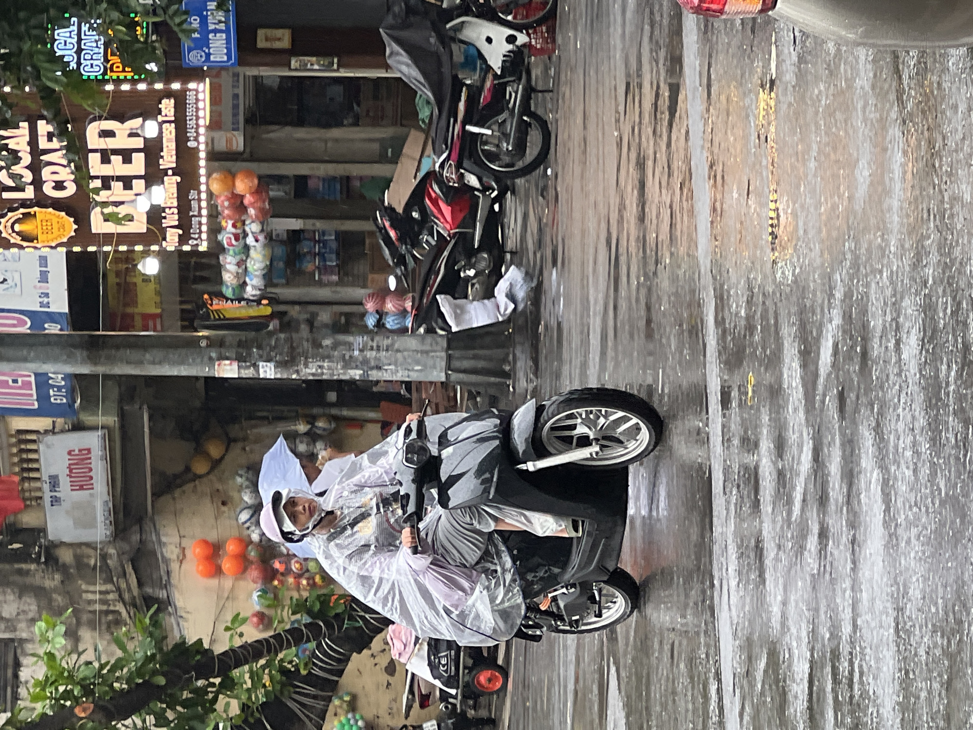 The image depicts a street scene in Hanoi, Vietnam, on a rainy day. A person is riding a black scooter, wearing a clear plastic rain poncho and a pink helmet. The street is wet and reflective, indicating recent rainfall. In the background, several shops are visible, including one with a prominent sign advertising "LOCAL CRAFT BEER" with the text "Tony Vu's Brewing - Vietnamese Taste" below it, along with an address and phone number. Other shops have signs with Vietnamese text. There are other motorbikes parked or in motion in the background. The overall atmosphere is that of a typical bustling street in Hanoi during a downpour.